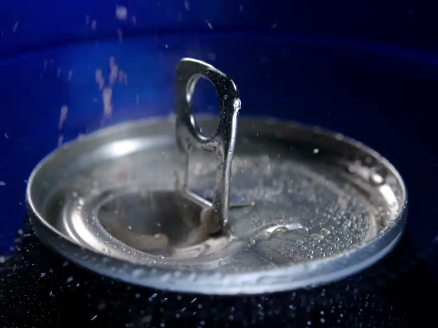 A close-up shot of a soda can being opened, with condensation and fizz visible on the metallic surface against a blue background.