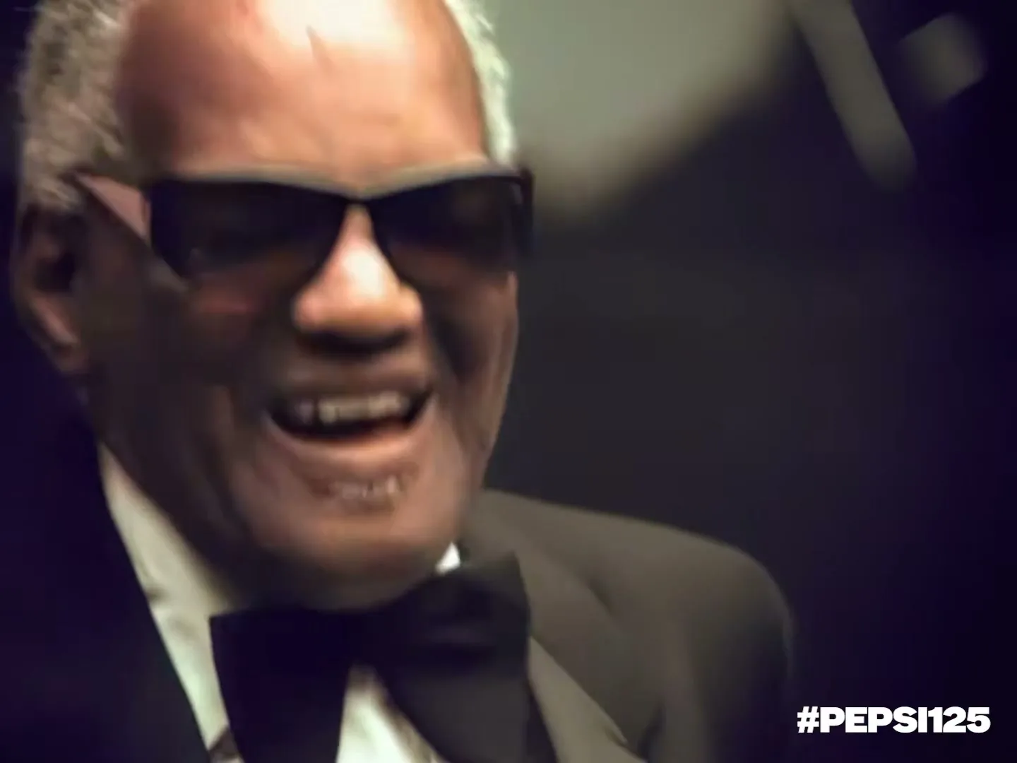 Ray Charles, an older Black man in a tuxedo and sunglasses, sings and smiles while playing the piano in a dark interior.