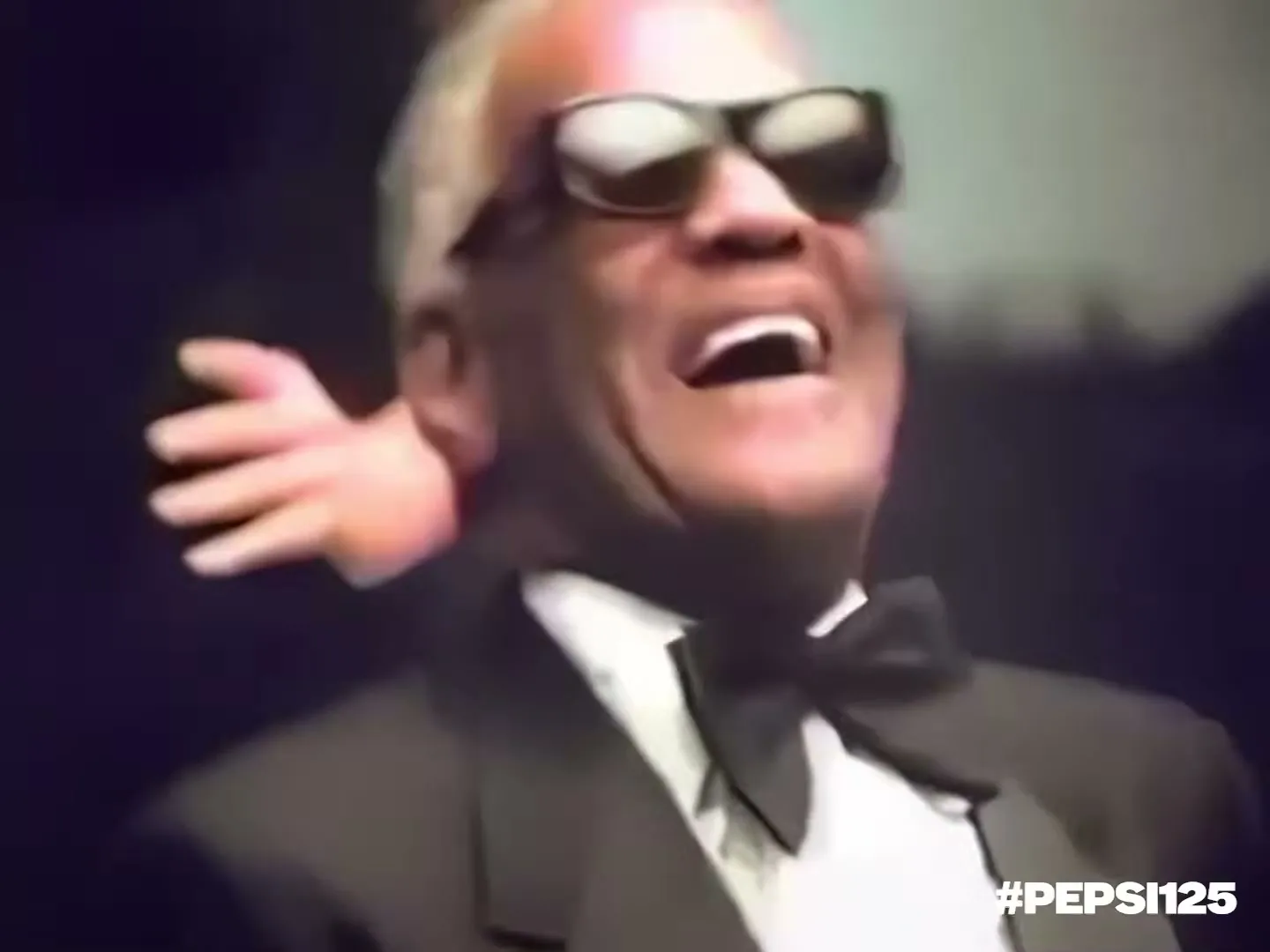 Ray Charles, a Black man in a tuxedo and sunglasses, laughs as a small doll hand appears to scratch his head.