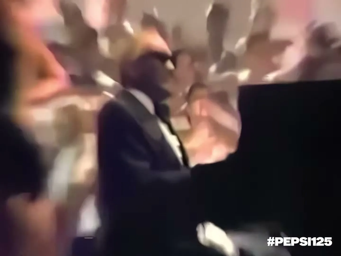 Ray Charles, a Black man in a suit and hat, plays piano on stage surrounded by dancing women in black dresses.