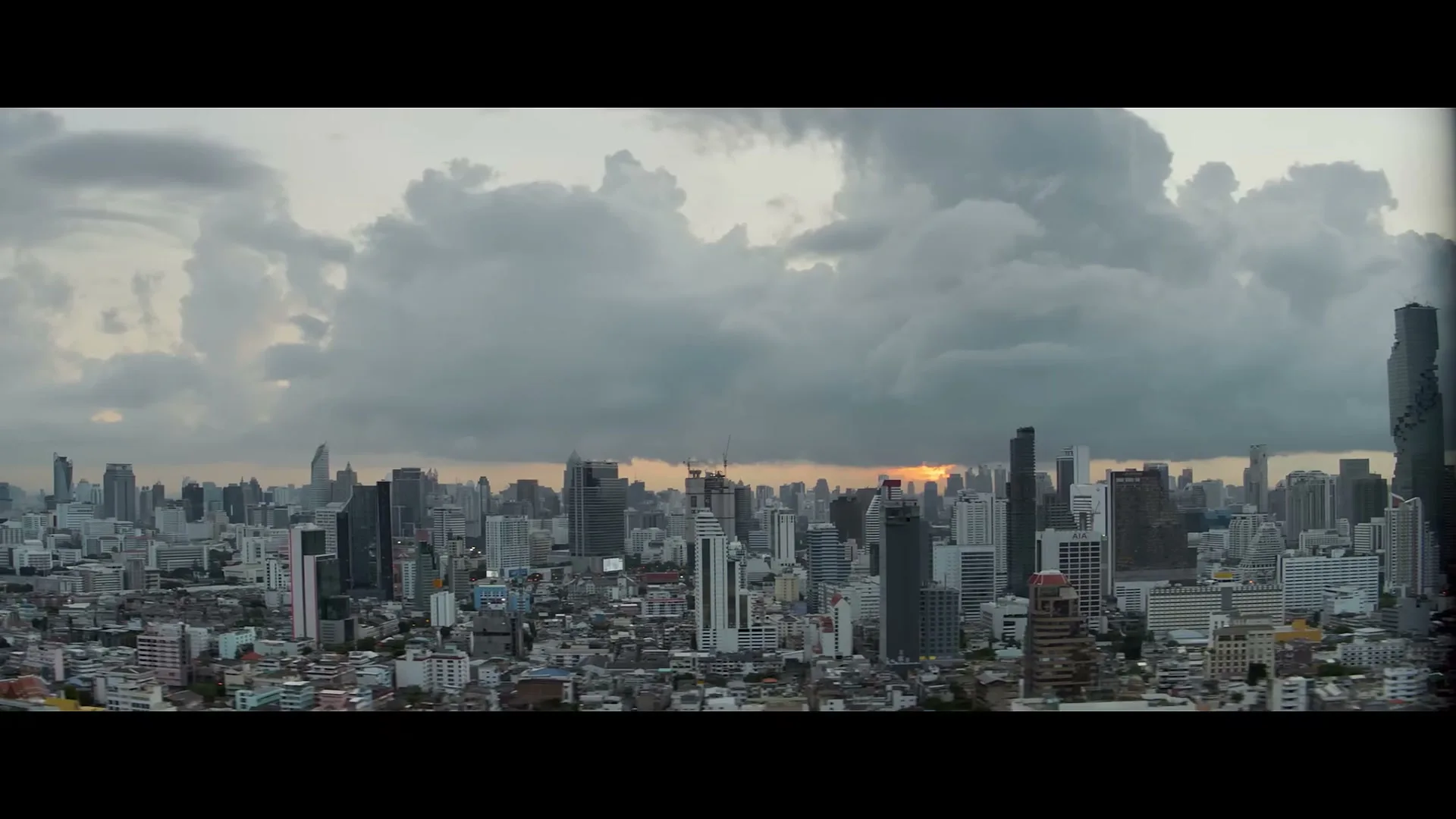 An establishing shot of a sprawling city skyline with numerous skyscrapers under a cloudy sky, with the sun setting on the horizon.