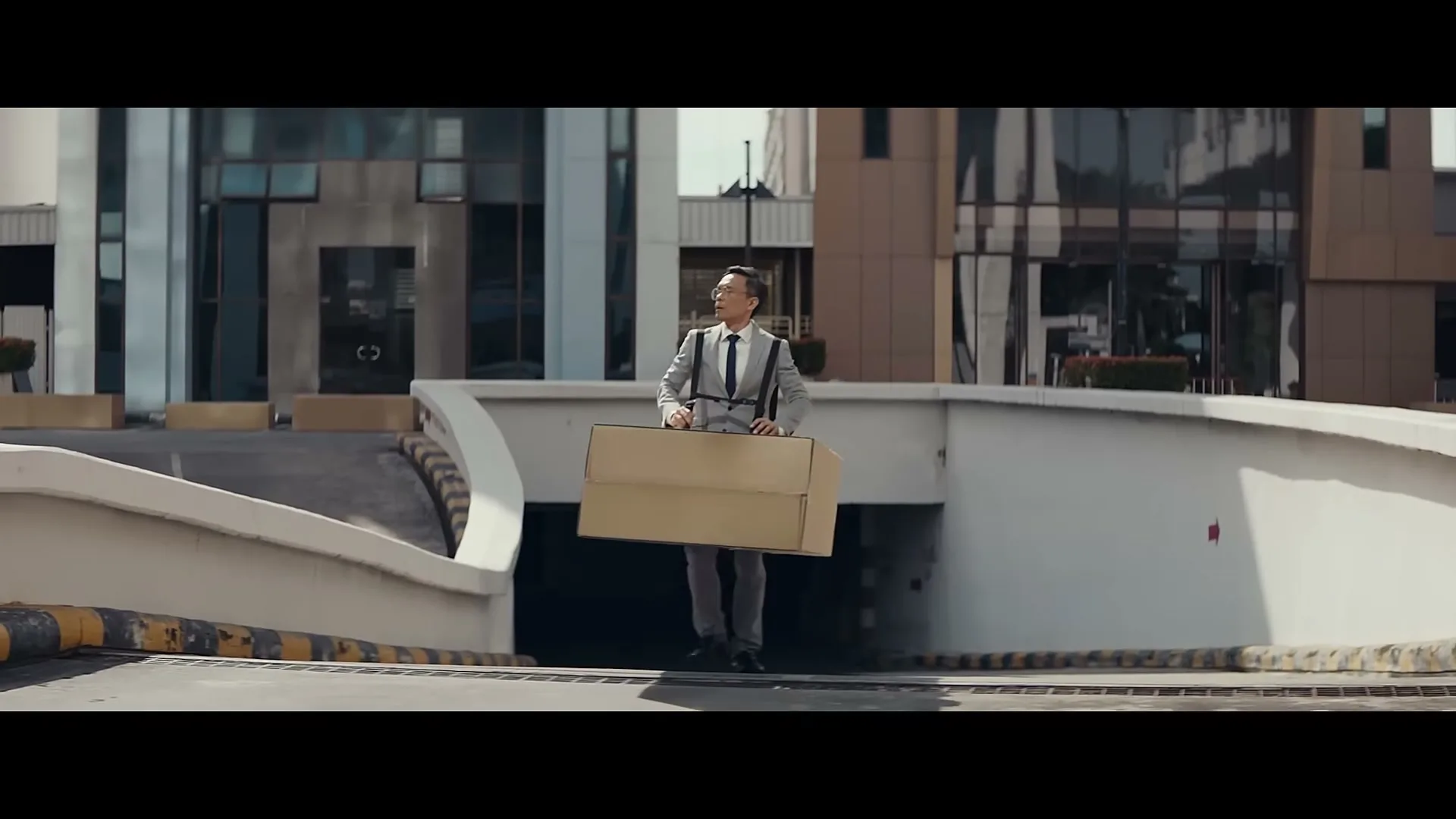 A man in a suit and suspenders, wearing a large cardboard box around his waist, walks out of a parking garage entrance on a sunny day.