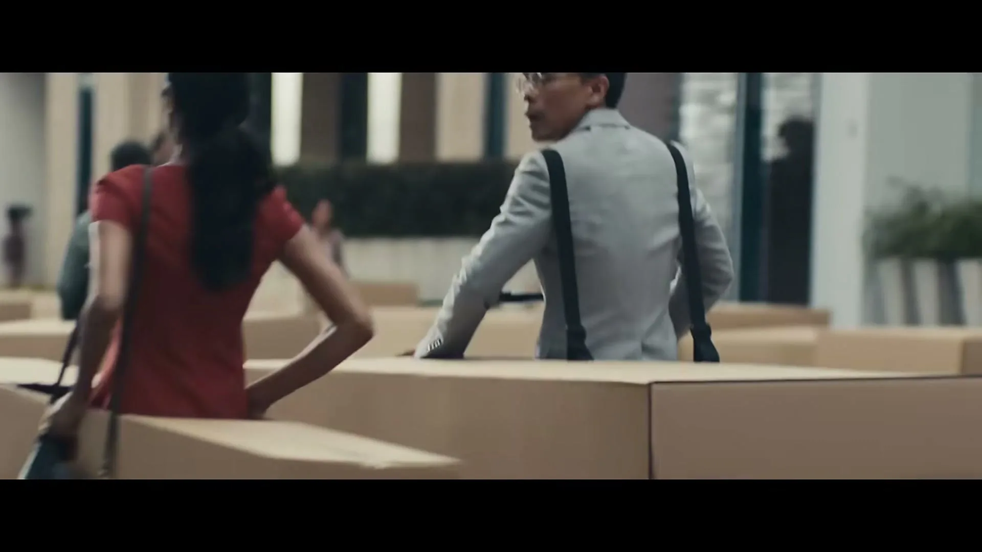 A man in a suit and suspenders looks around a maze of cardboard boxes, while a woman in a red shirt walks past him.