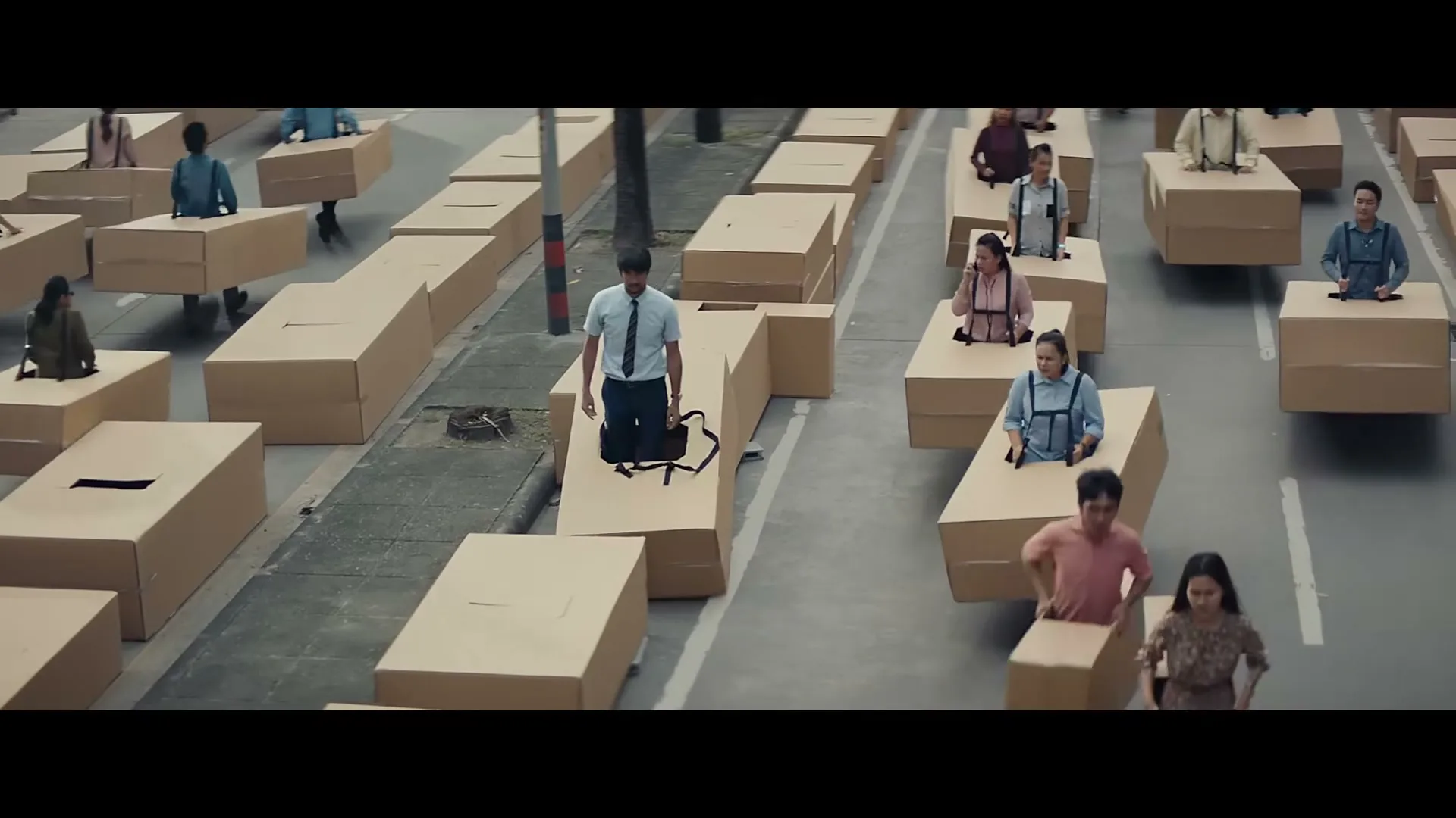 A man in a light blue shirt and dark tie stands on a cardboard box, surrounded by many other people also in cardboard boxes, moving along a street.