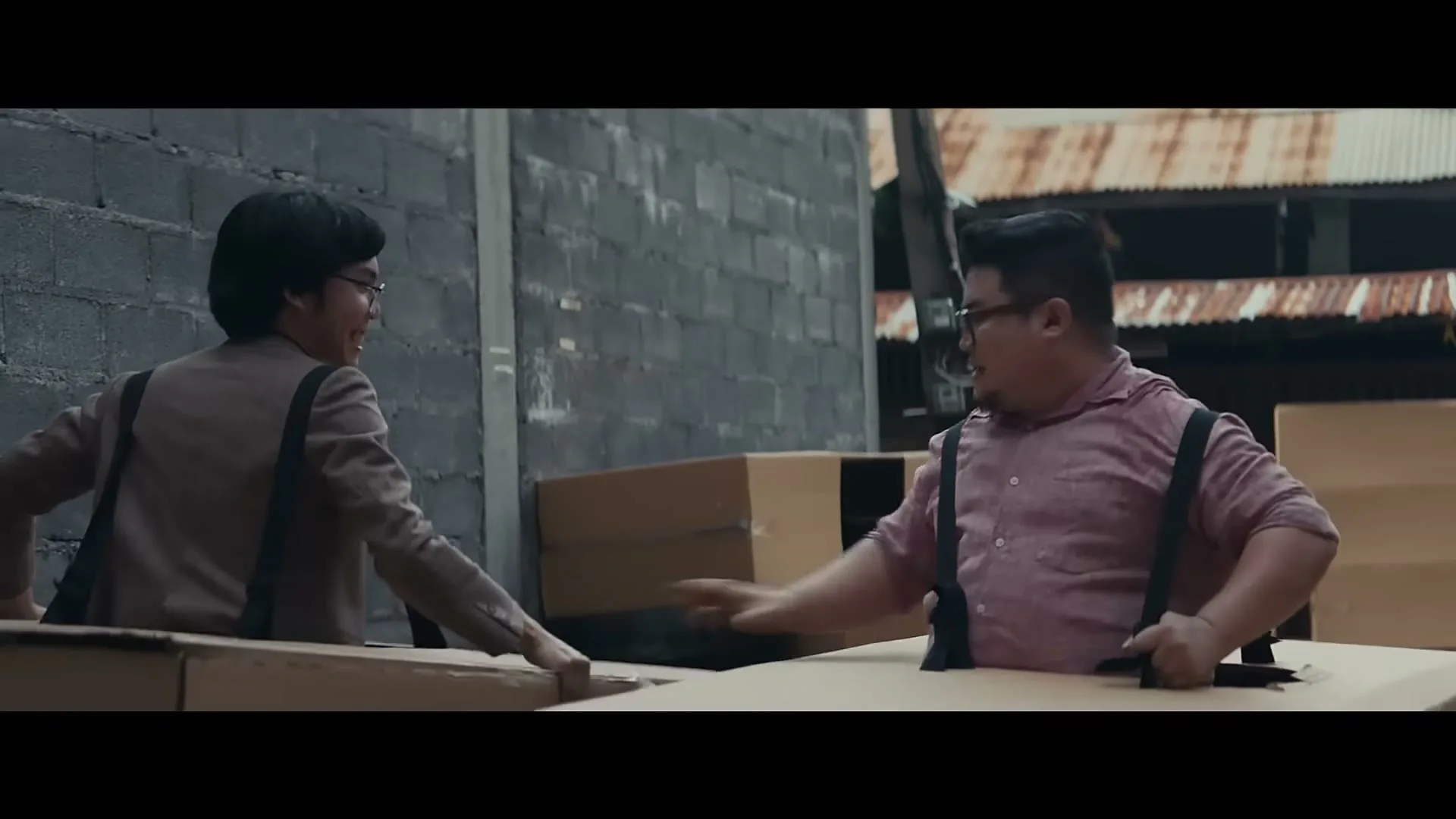 Two men, one in a brown jacket and the other in a pink shirt with suspenders, are playing in cardboard boxes in an alleyway.