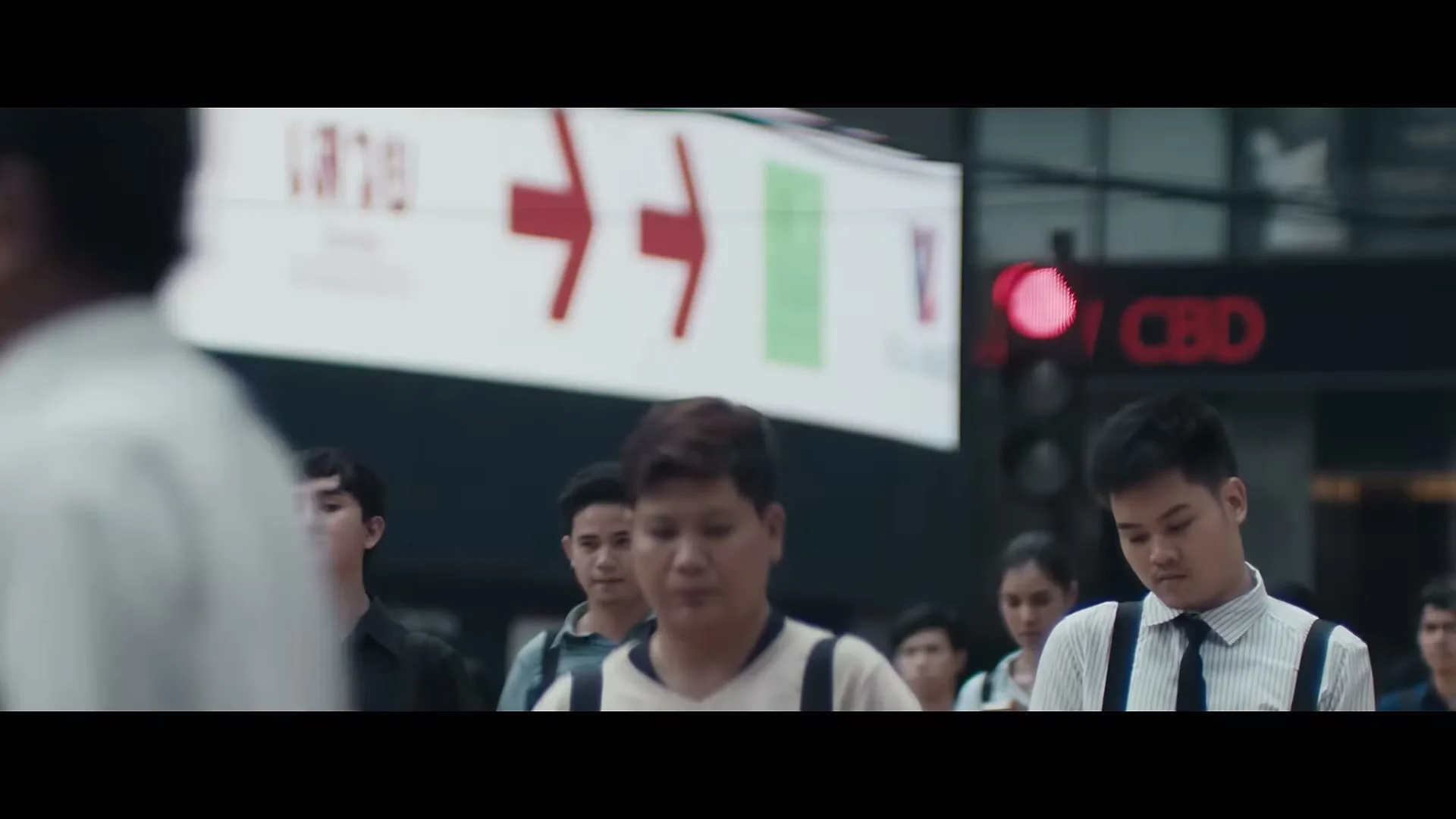 A diverse crowd of people walks on a busy city street, with a young man in a white shirt and suspenders looking down, and a large sign with red arrows in the background.