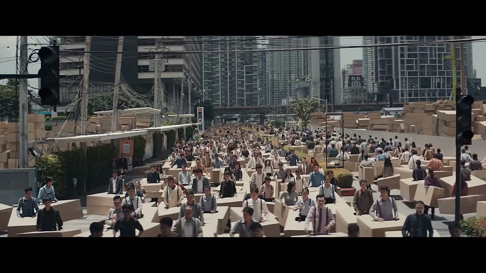 A wide shot shows a large crowd of people, mostly men, walking through a city street, each encased in a cardboard box, under a bright sky.