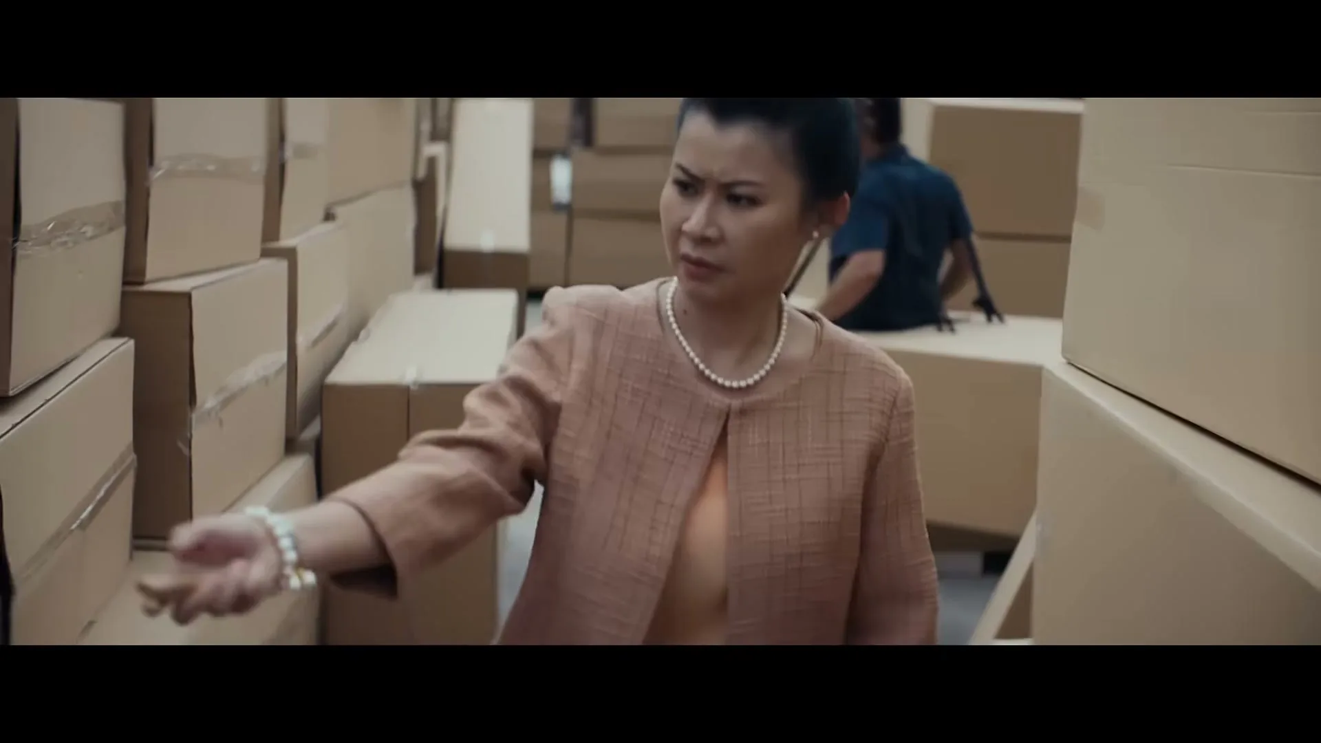 A woman in a peach suit and pearl necklace looks around with a confused expression, surrounded by stacks of cardboard boxes in a warehouse.