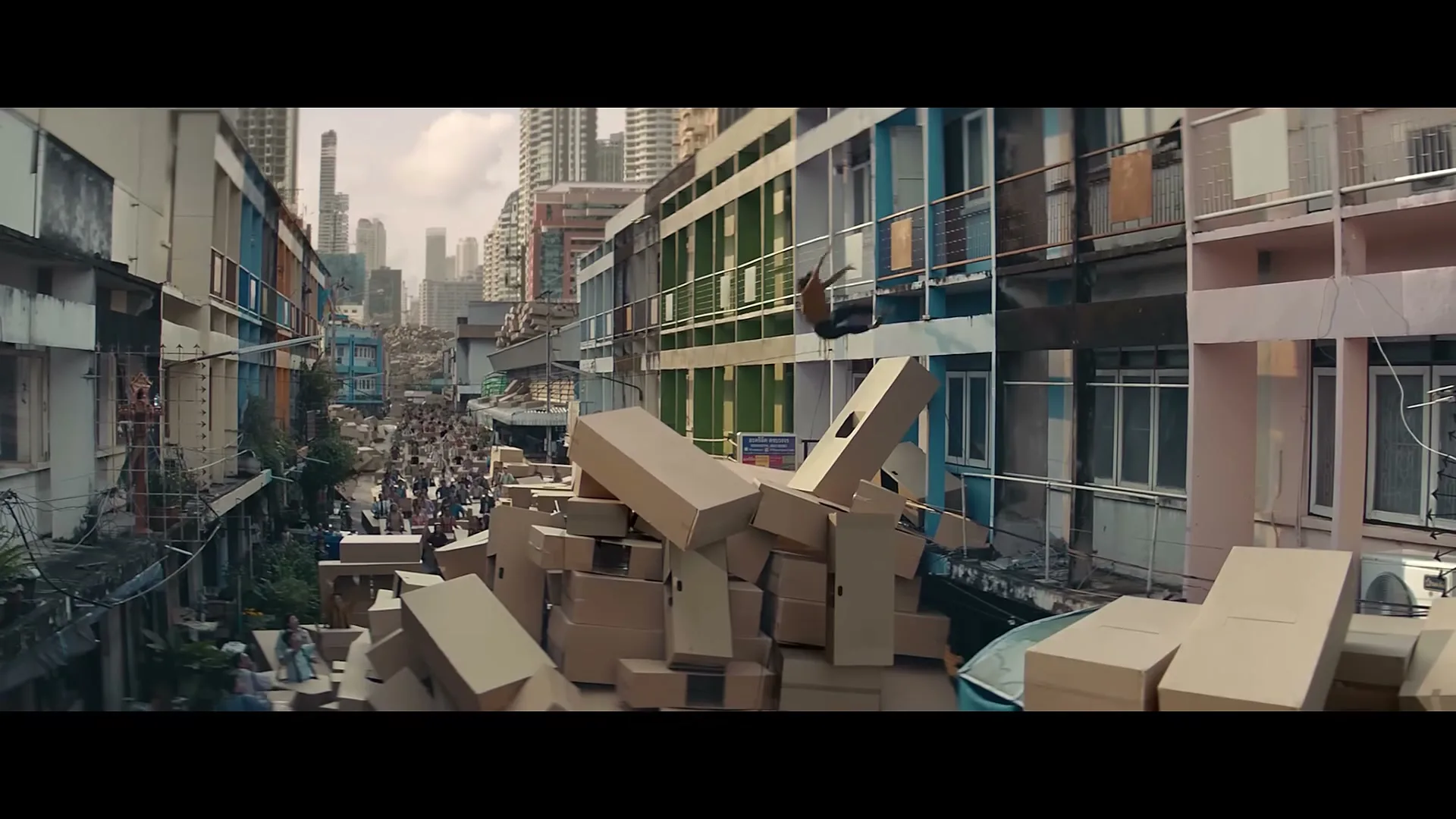 A man in a yellow shirt and dark pants falls from a balcony onto a pile of cardboard boxes in a crowded city street.