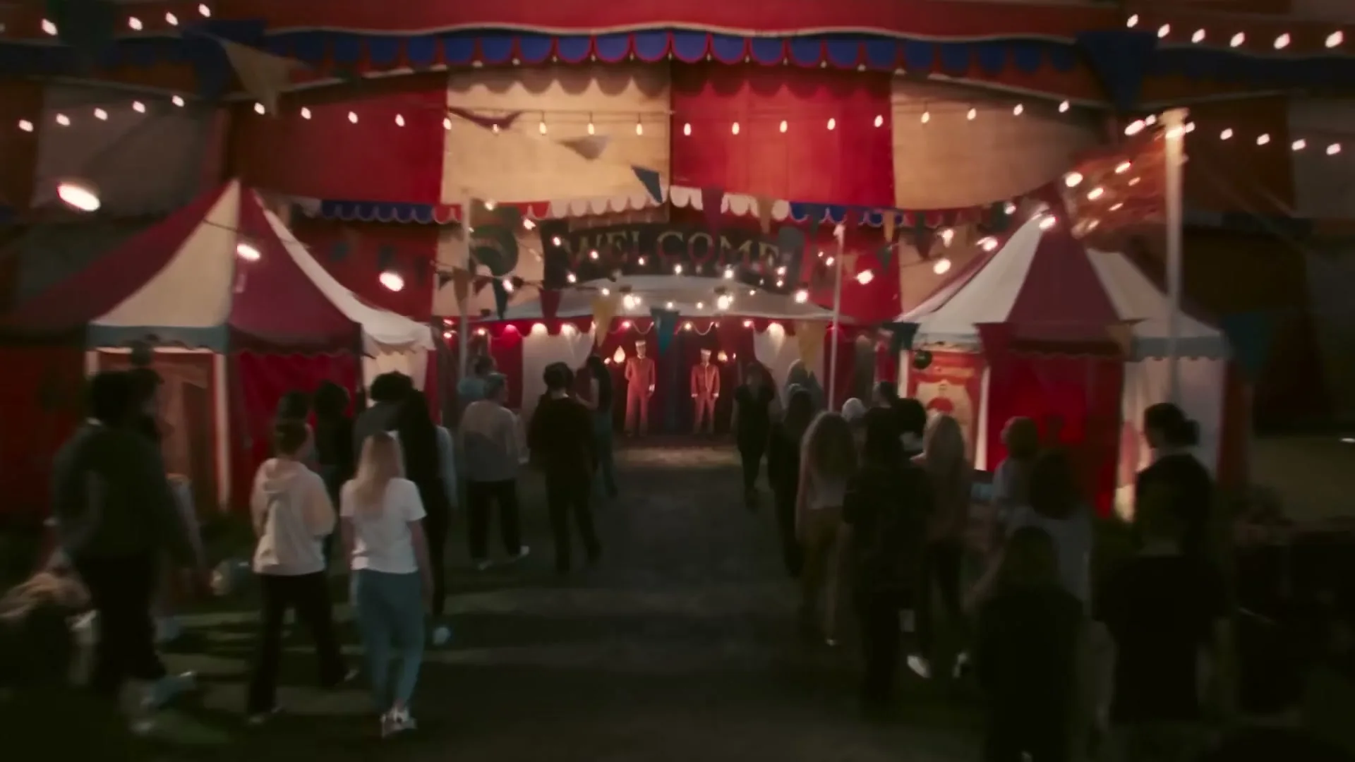 A large, red and white striped circus tent with many lights is seen at night, as people walk towards its entrance.