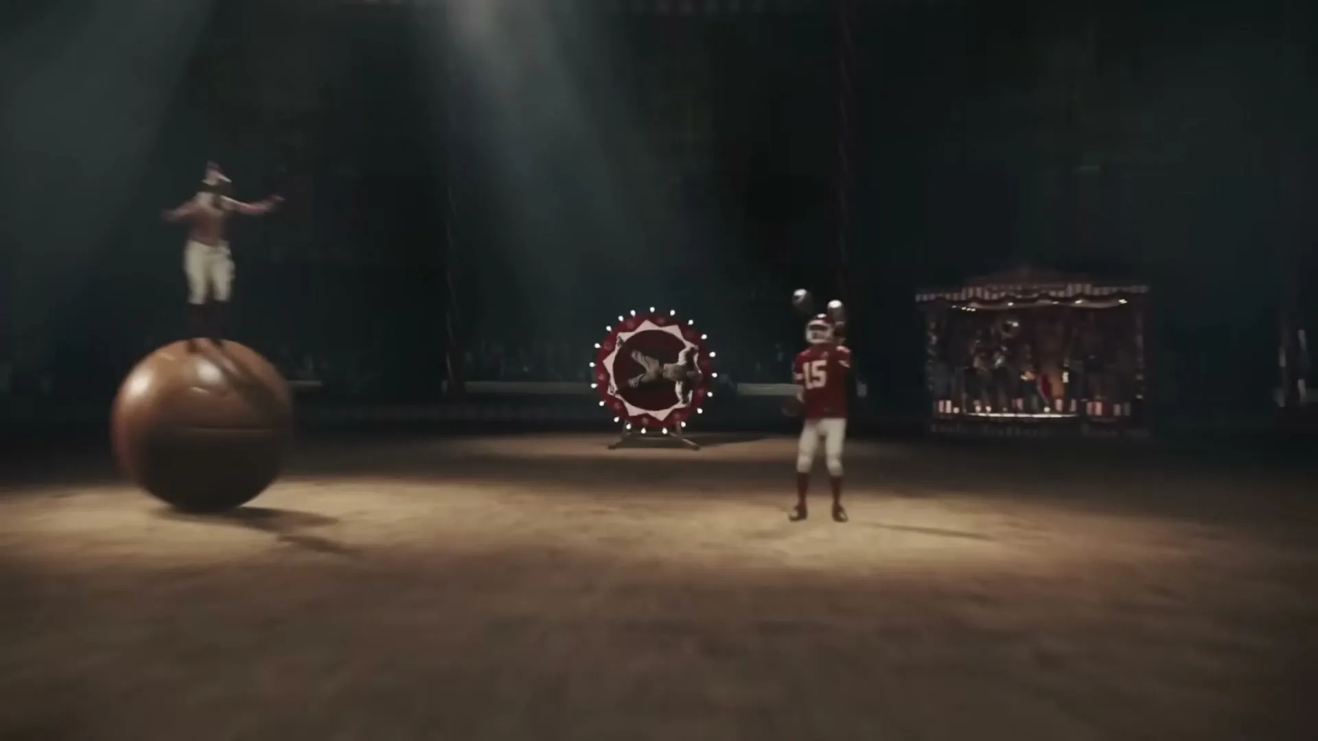 A man in a vintage circus costume is strapped to a spinning wheel with lights, as the camera pulls back to reveal a circus ring with various performers.