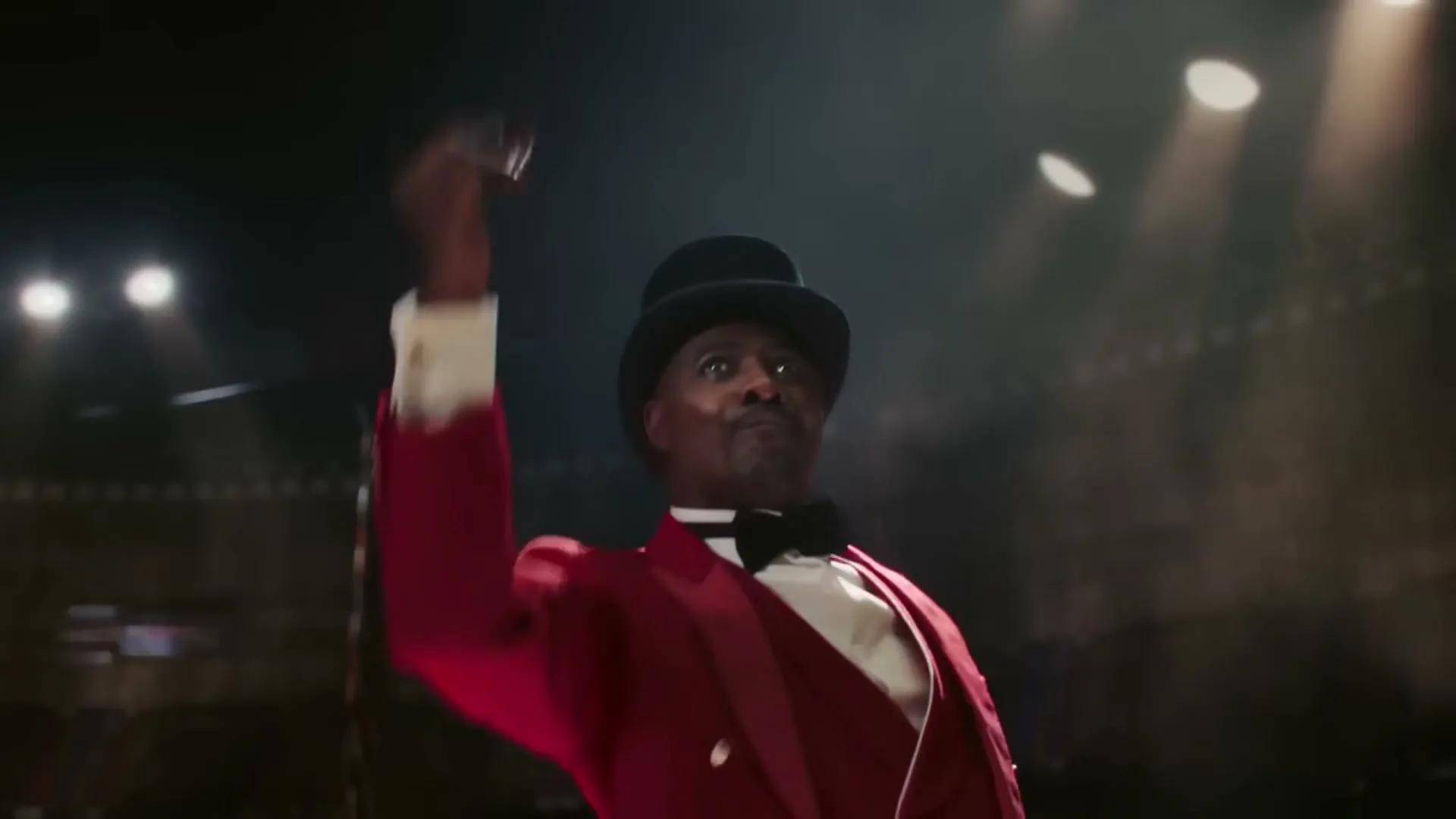 A Black man in a red ringmaster suit and top hat raises his arm in a spotlight, looking up in a stadium.