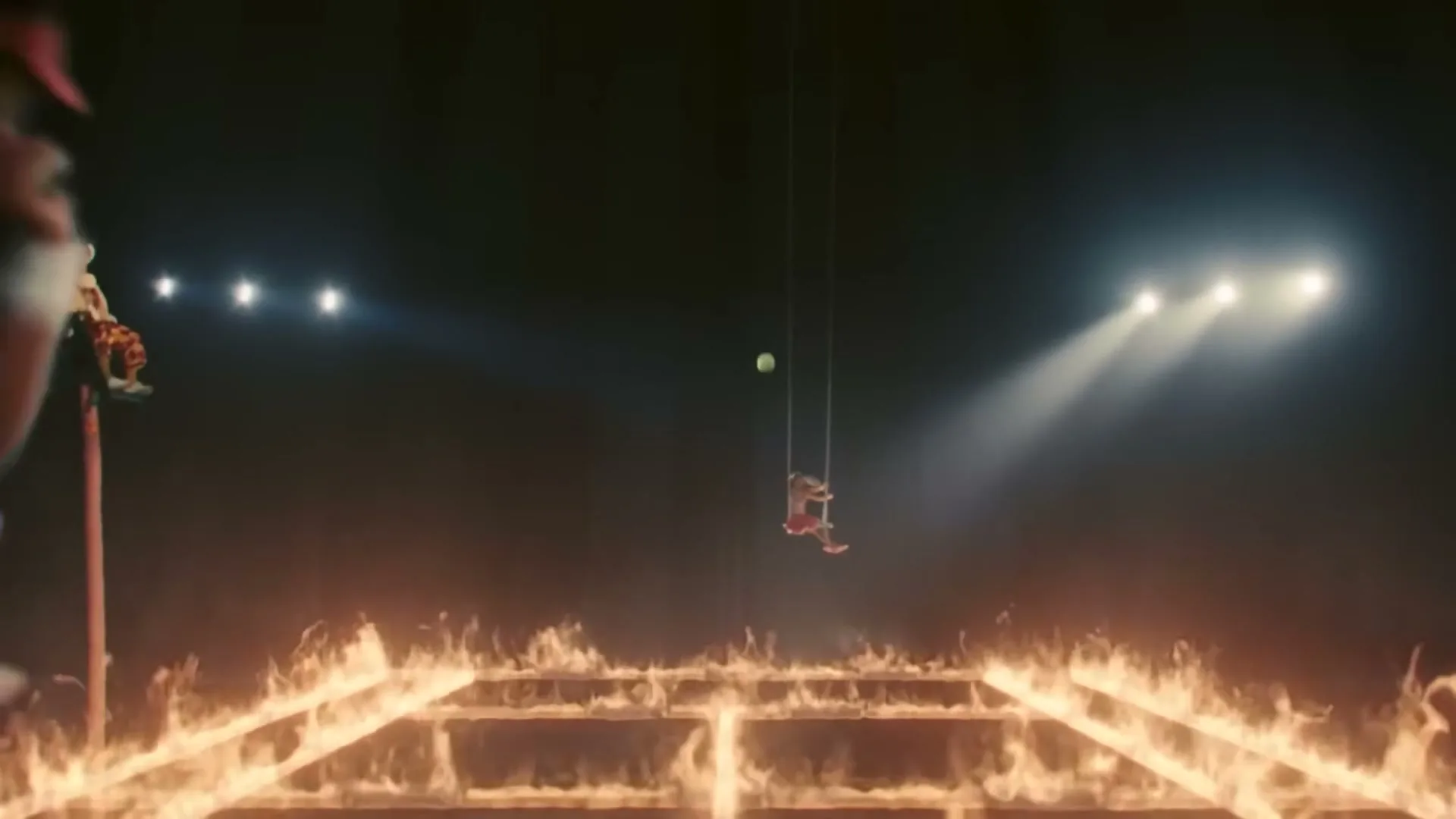 A tennis player swings on a trapeze, hitting a tennis ball over a flaming tennis court, while another performer balances on a pole.