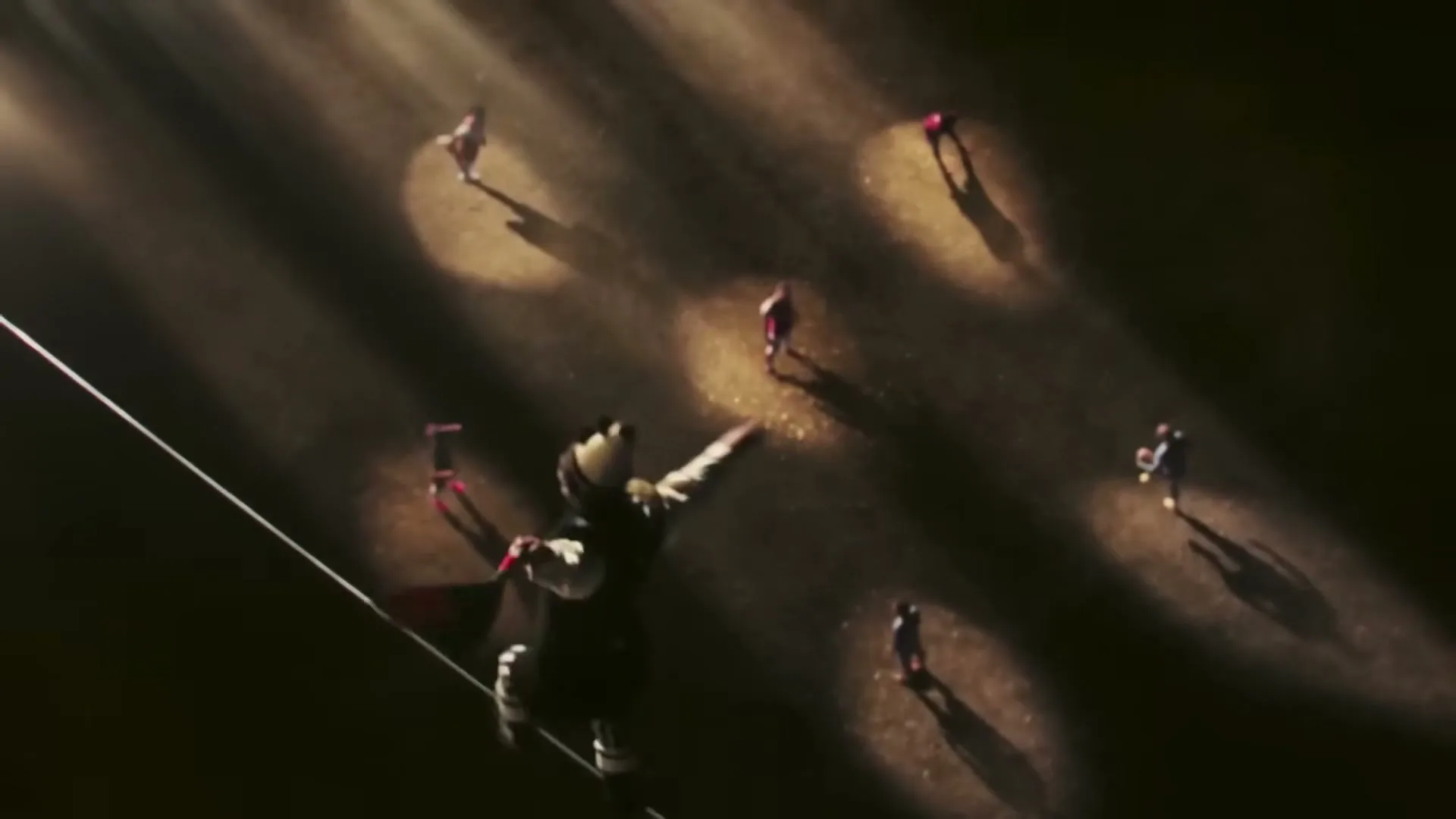 A male tightrope walker in a circus costume balances on a rope above a dark ground with multiple spotlights on other people.