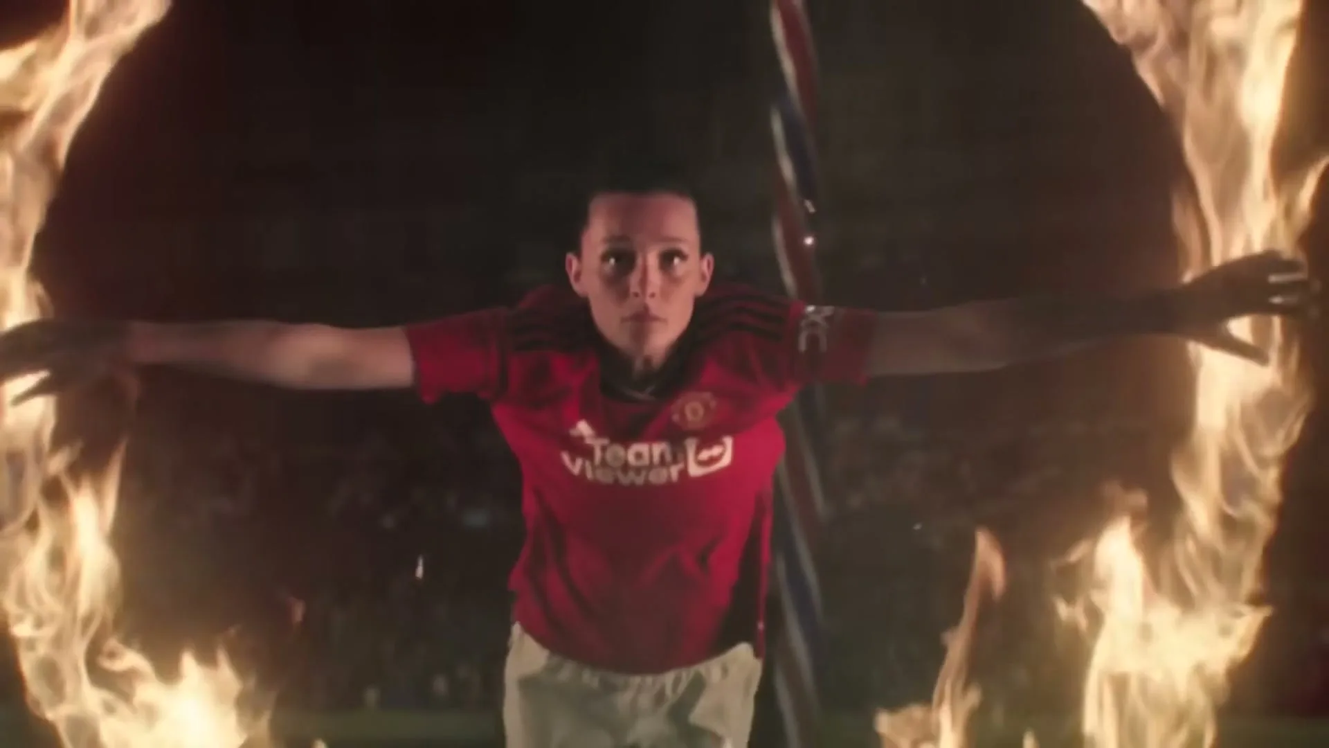 A female soccer player in a red jersey stands with arms outstretched, surrounded by flames in a dark stadium.