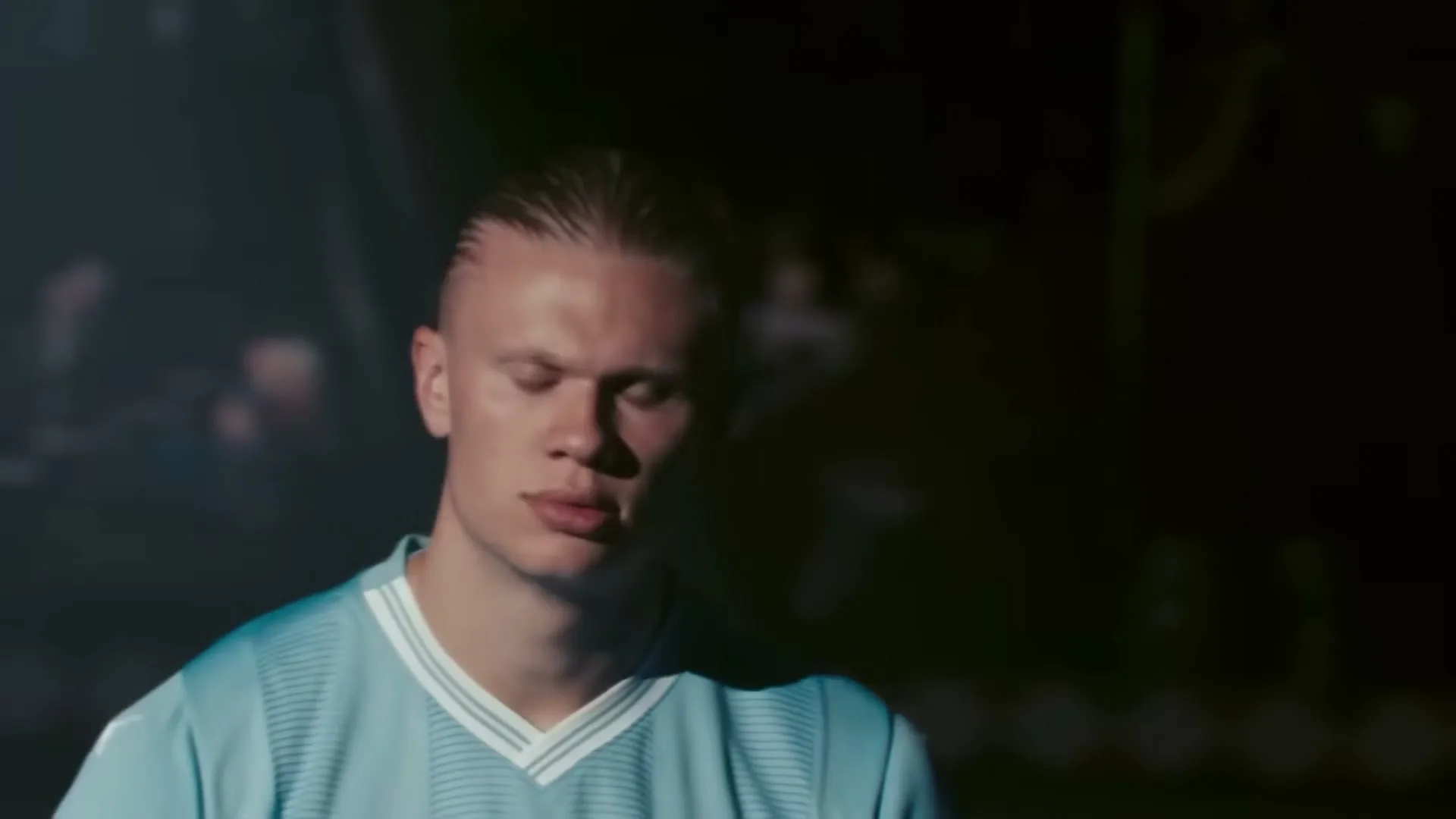 A male footballer in a light blue jersey looks directly at the camera, then closes his eyes, then looks down, in a dark stadium tunnel.