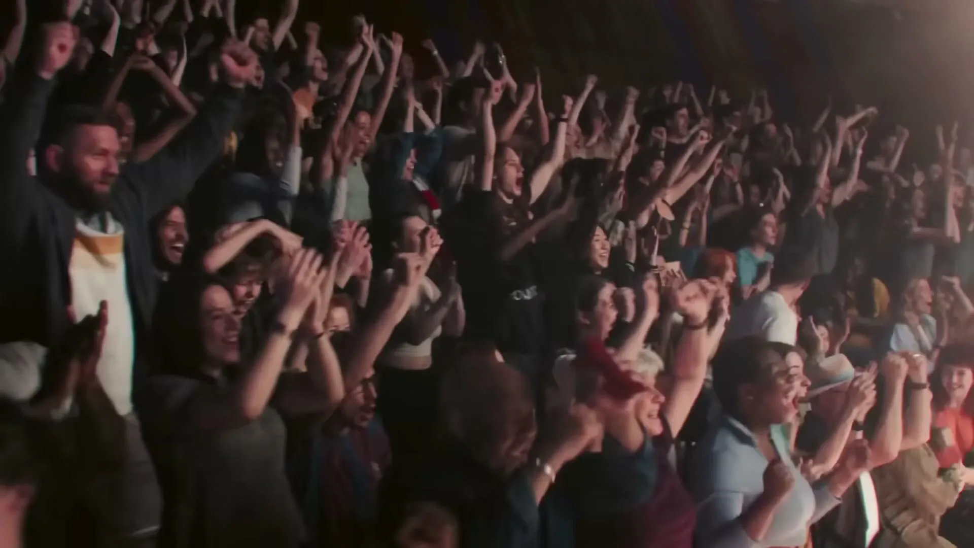 A diverse crowd of people cheers with their arms raised in a dimly lit stadium or arena, celebrating an unseen event.
