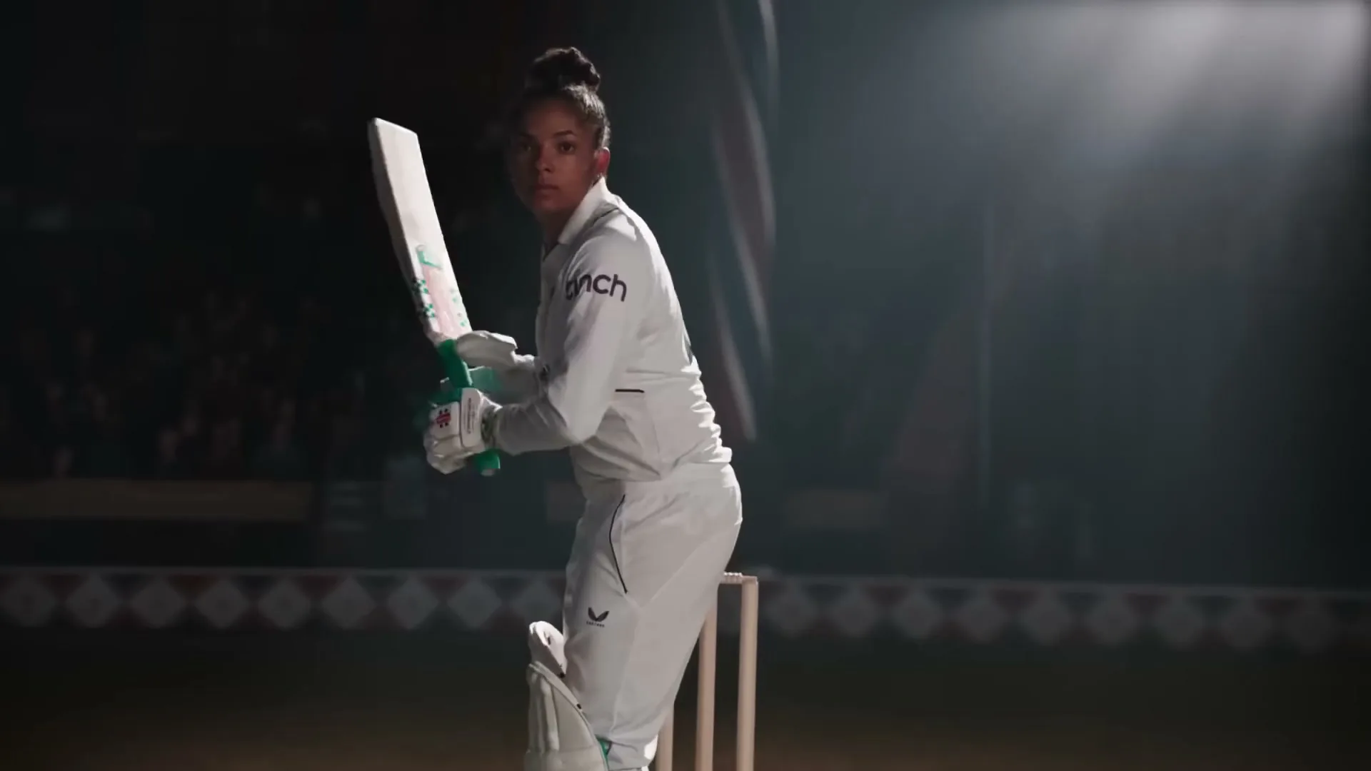 A female cricket player in white uniform holds a bat, looking intently at the camera in a dimly lit stadium.
