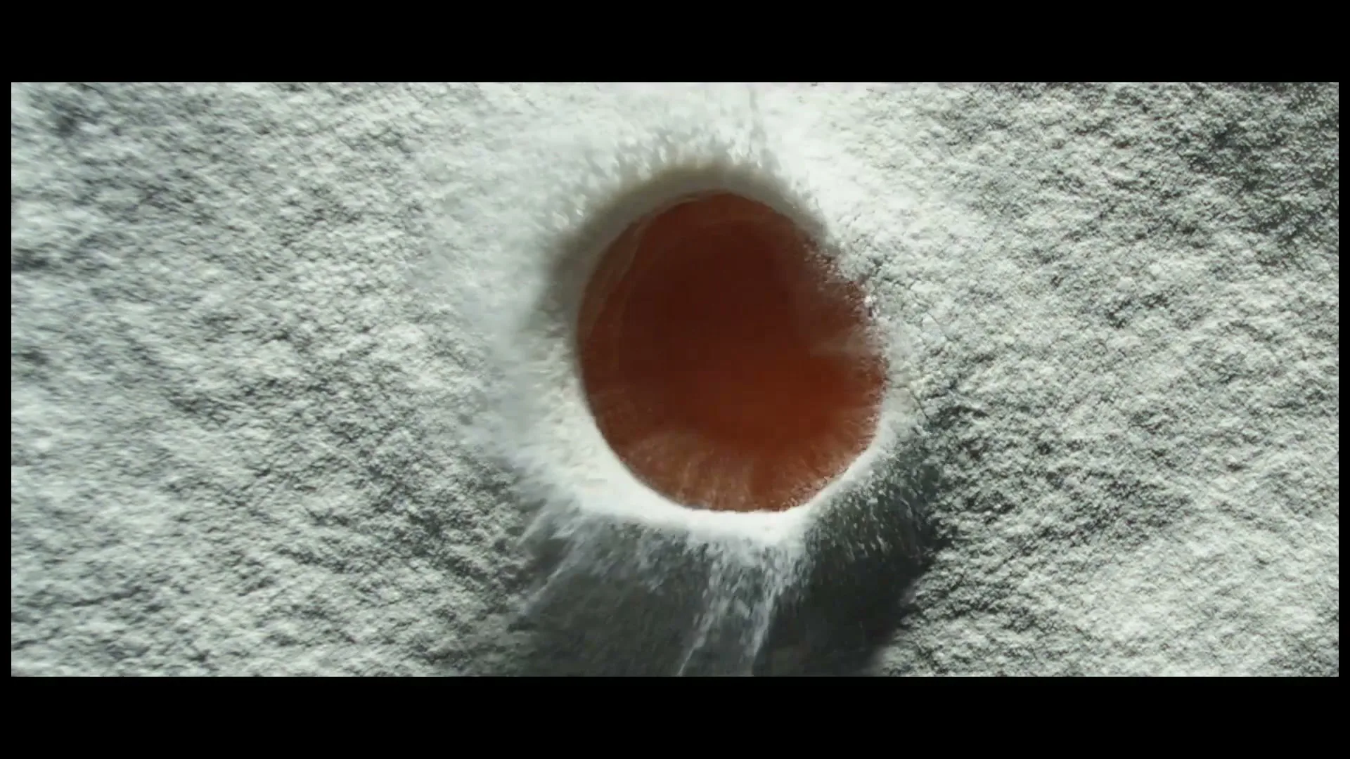 A single, shiny orange egg yolk is dropped into a bed of white flour, creating a small crater and splashing flour upwards.