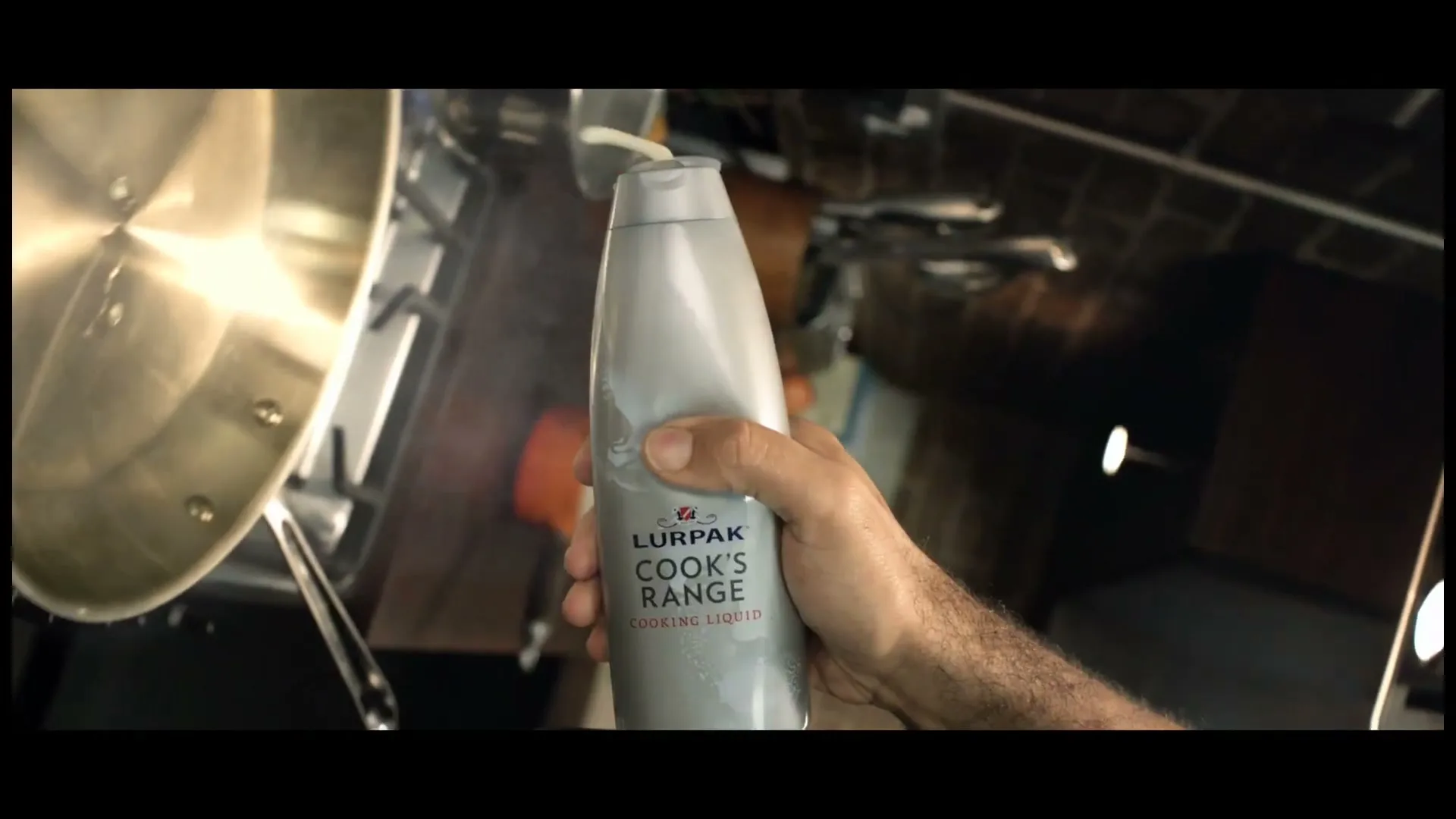 A male hand holds a silver Lurpak Cook's Range Cooking Liquid bottle, squeezing it to dispense liquid into a pan on a stove in a kitchen.