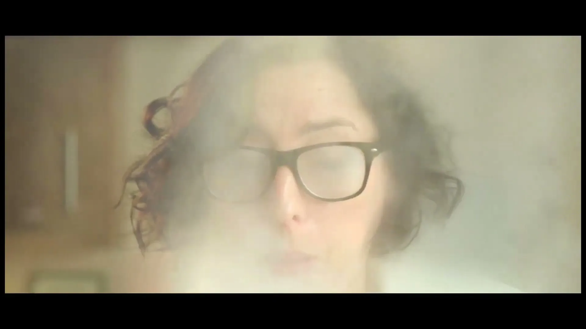 A woman with dark, curly hair and black-rimmed glasses blows steam from a hot dish, fogging her glasses in a kitchen.