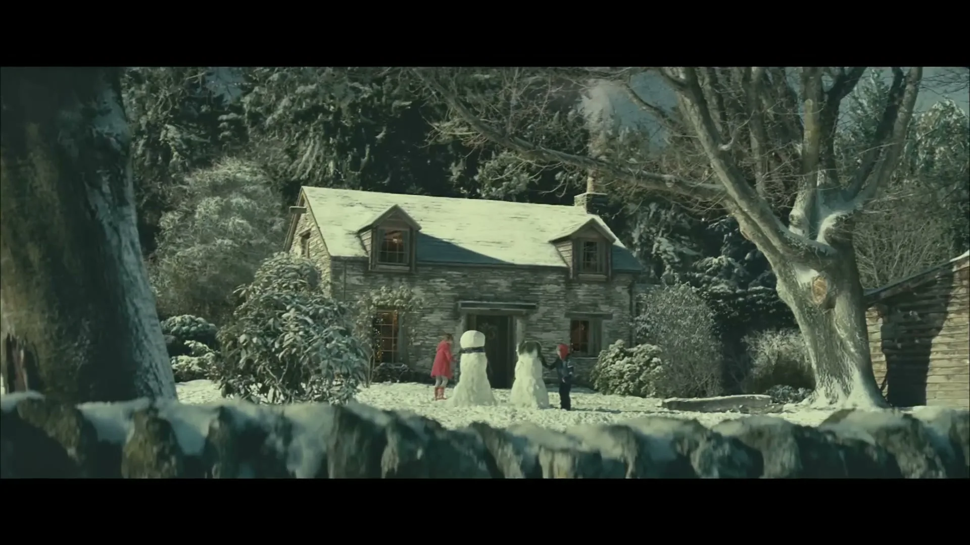 A wide shot of a snowy cottage with two snowmen and two people standing outside, surrounded by snow-covered trees and a stone wall.