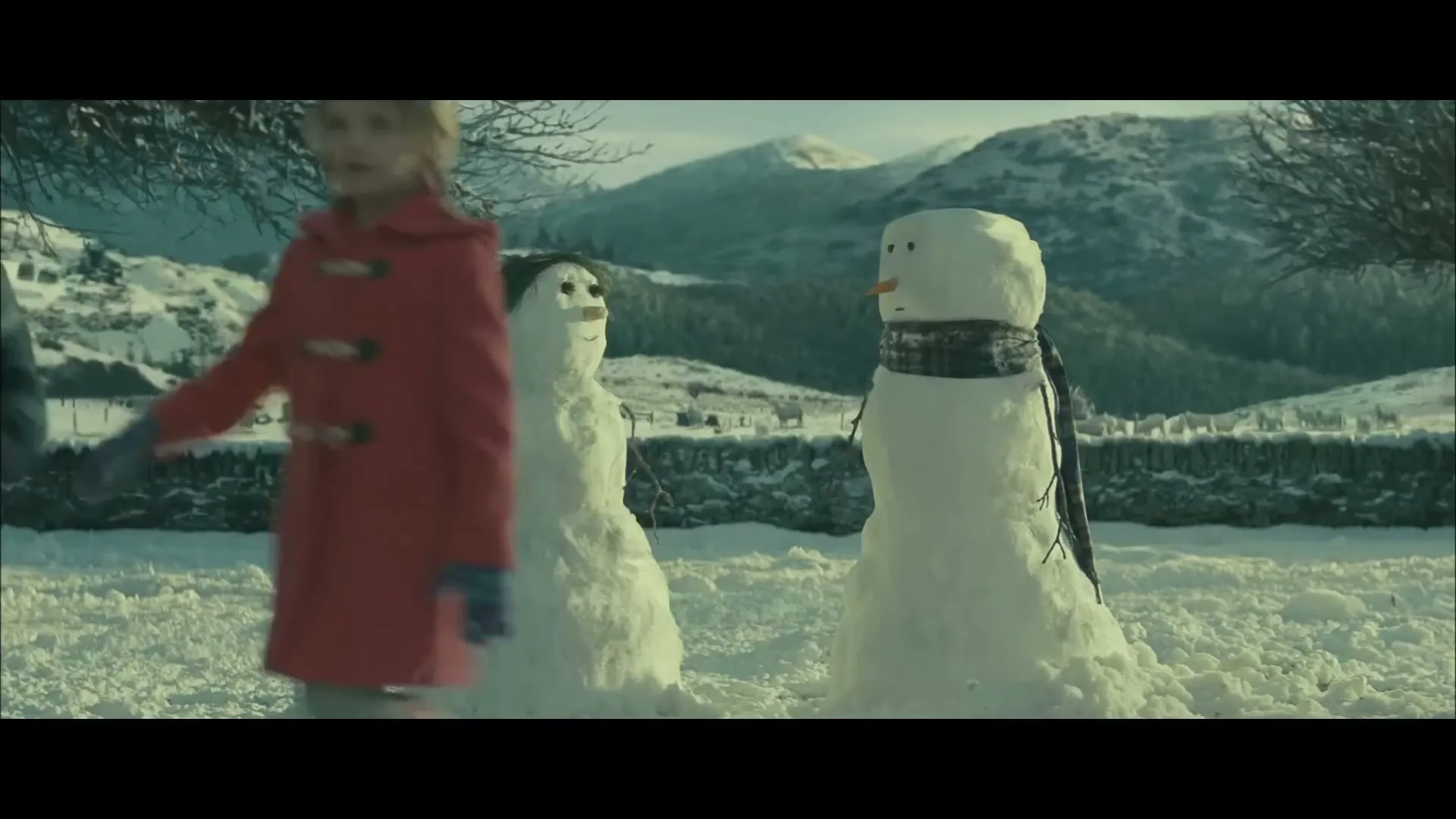 Two children, a boy and a girl, walk past two snowmen in a snowy, mountainous landscape during the day.