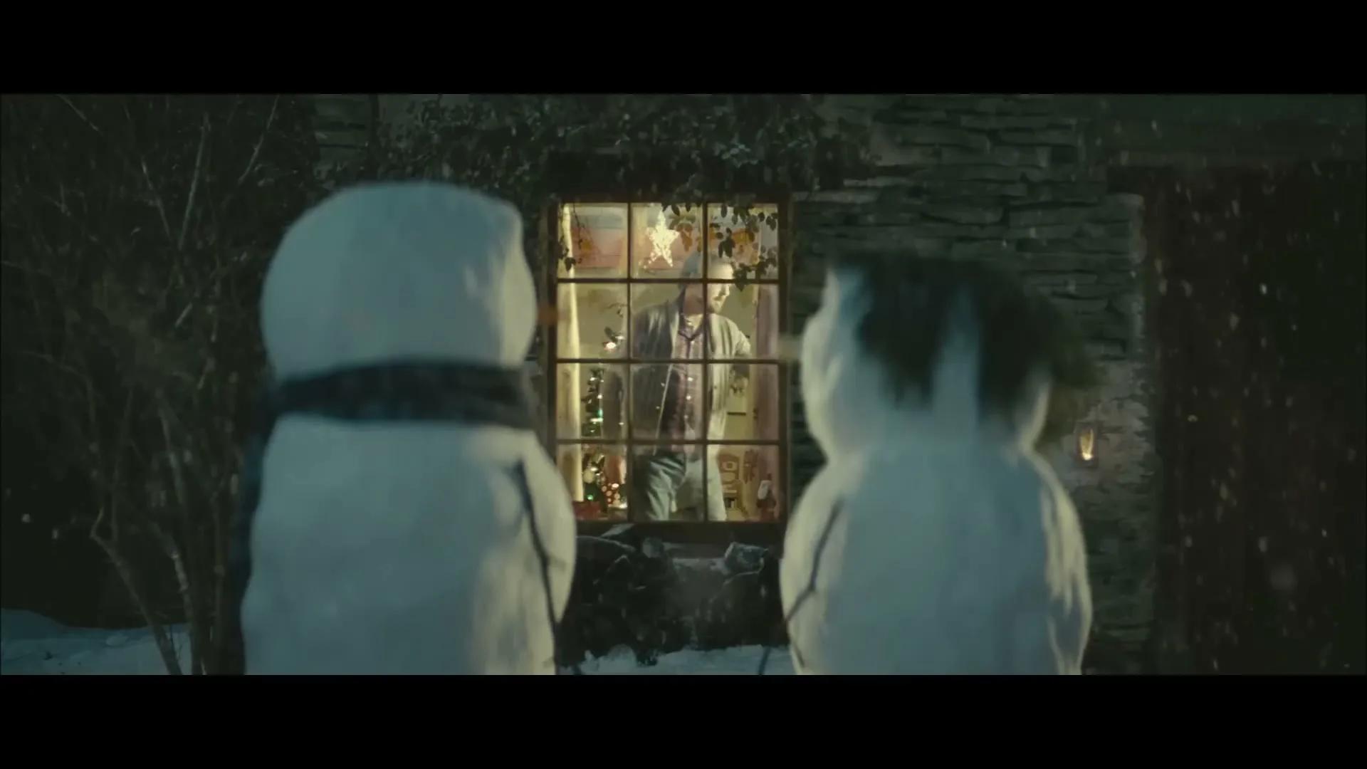 Two snowmen stand outside a house at night, looking through a window at a man inside who is decorating a Christmas tree.
