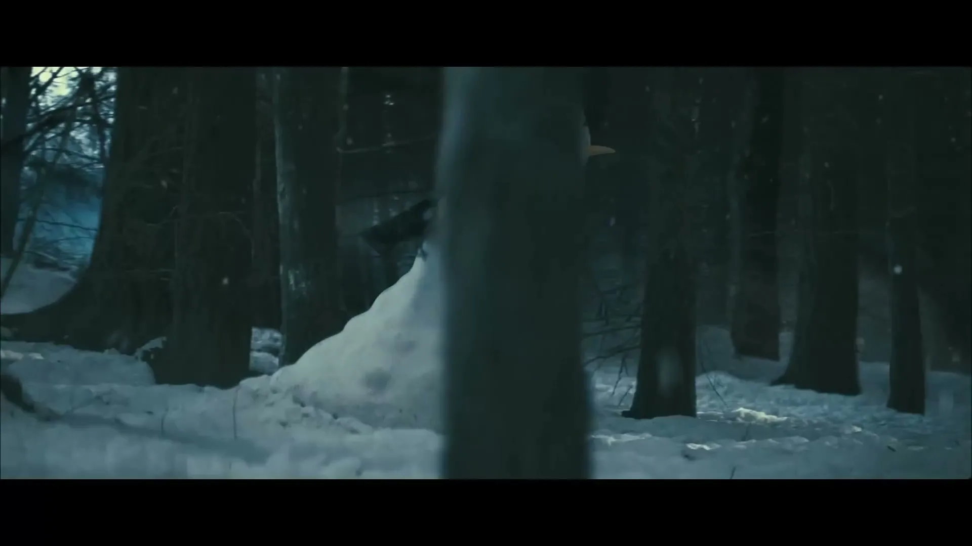 A snowman with a scarf and carrot nose stands in a snowy forest, partially obscured by trees, as the camera slowly pans right.