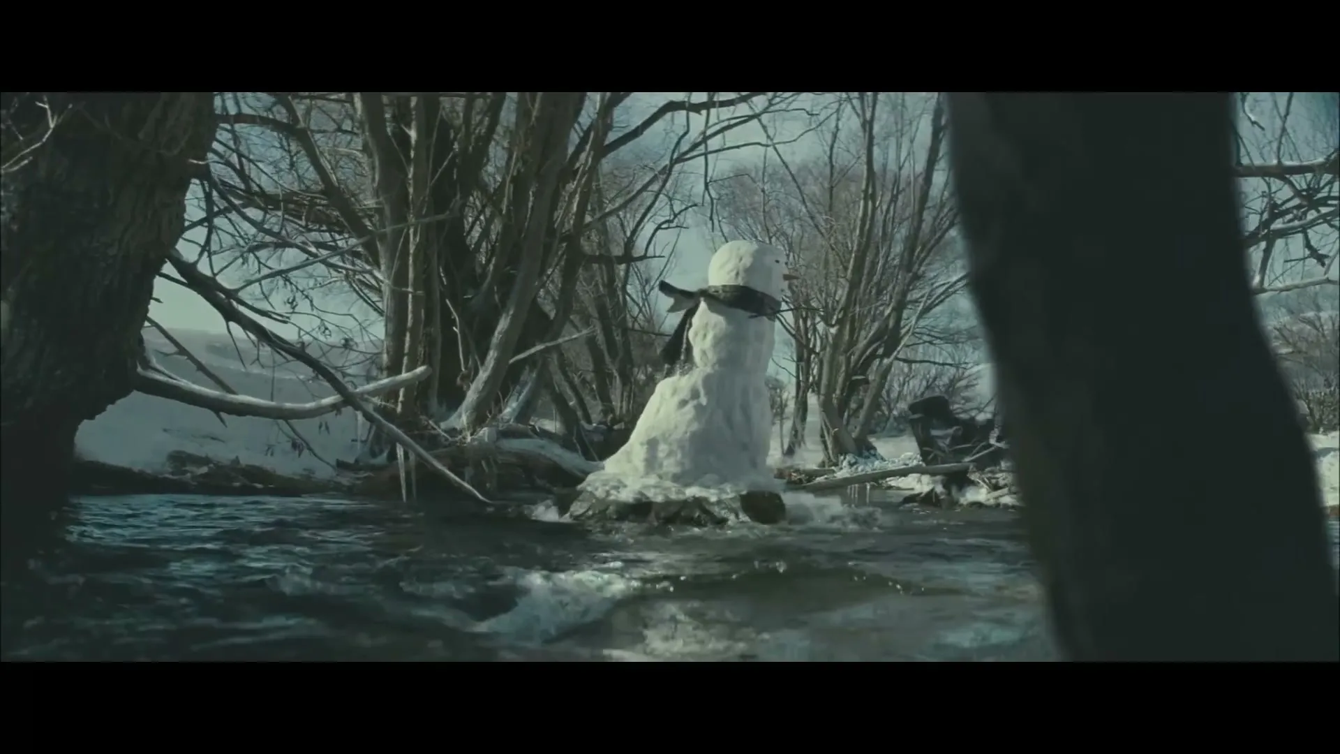 A snowman with a scarf stands in a flowing river, surrounded by snow-covered trees and banks in a winter landscape.