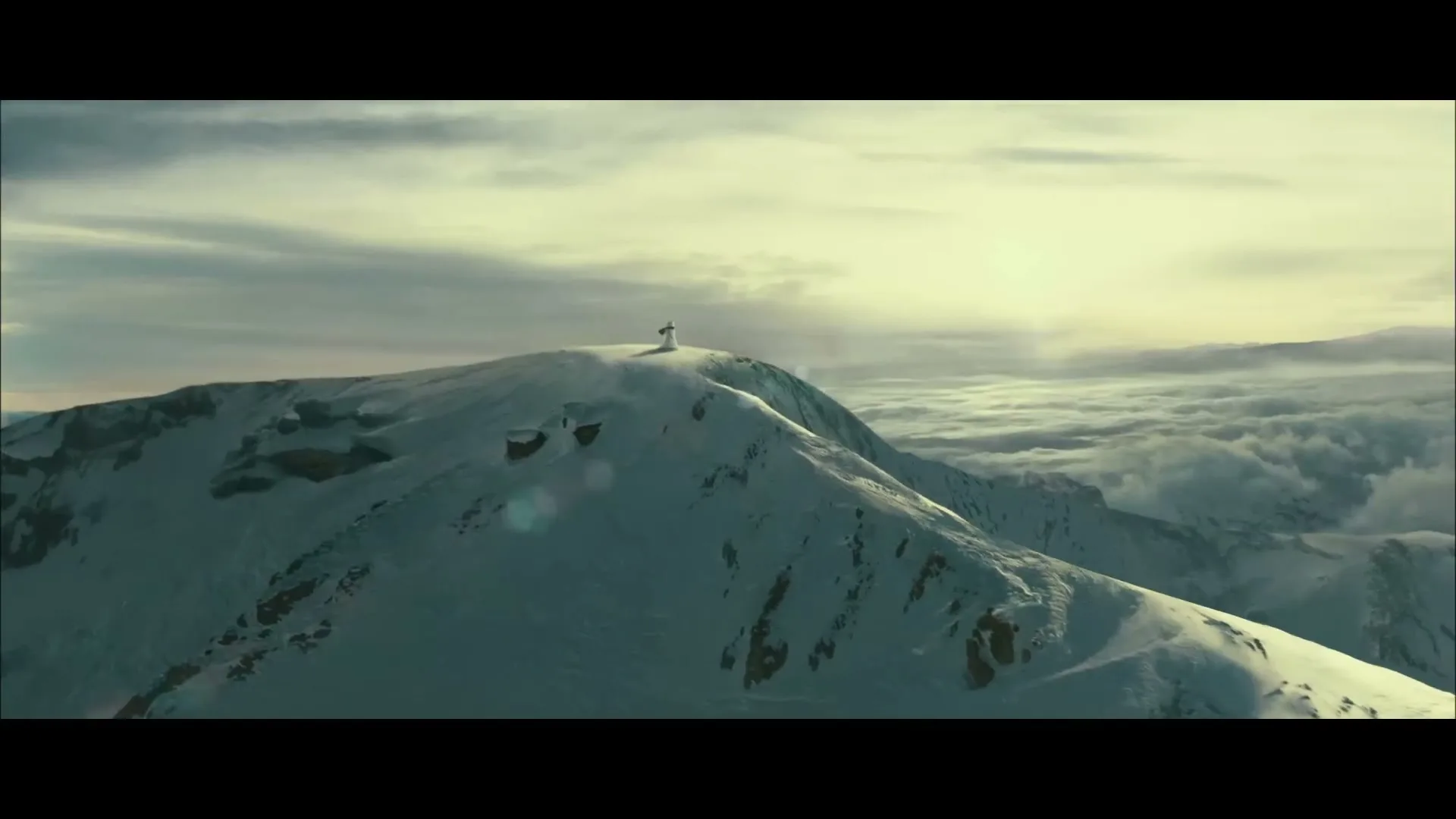 A lone snowman stands atop a snow-covered mountain peak, with clouds stretching out below and a bright sky above.