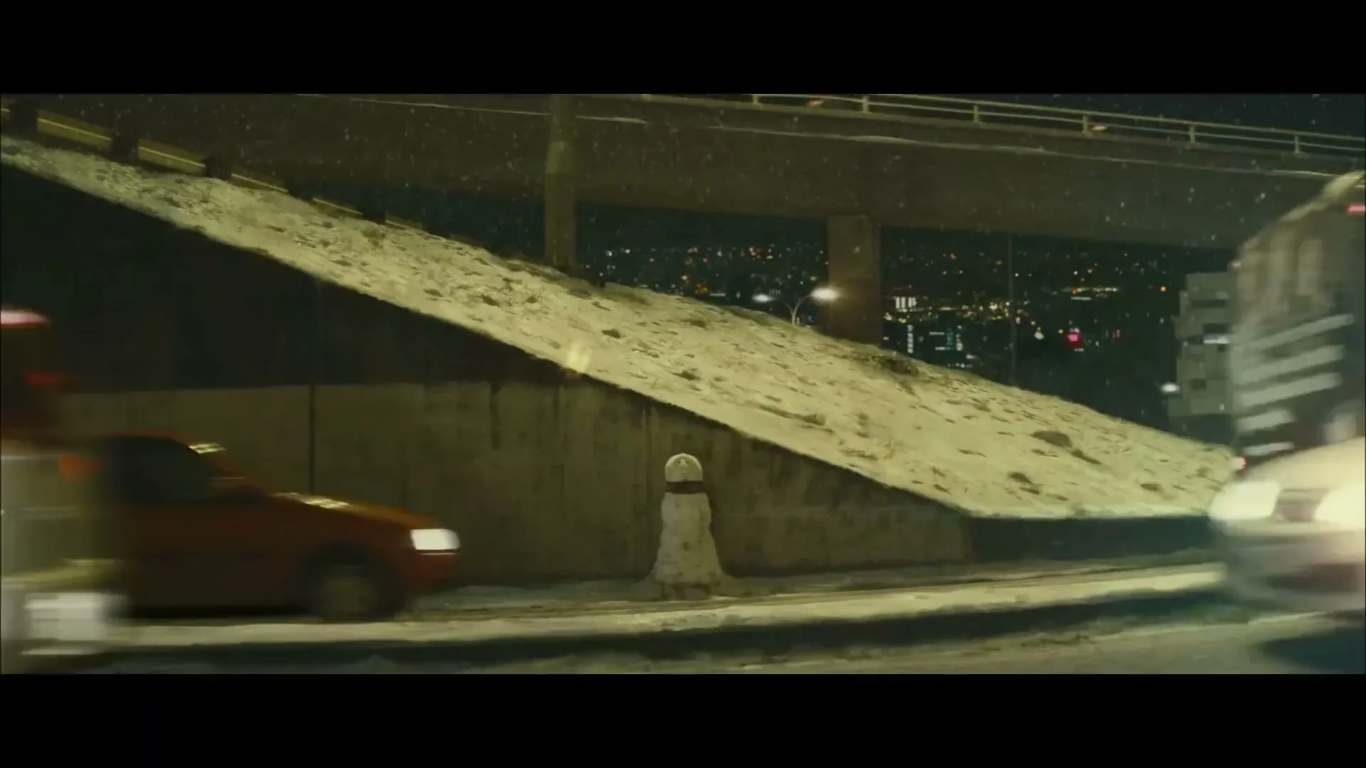 Cars and a bus drive past a snow-covered embankment next to a highway overpass at night, with city lights in the background.