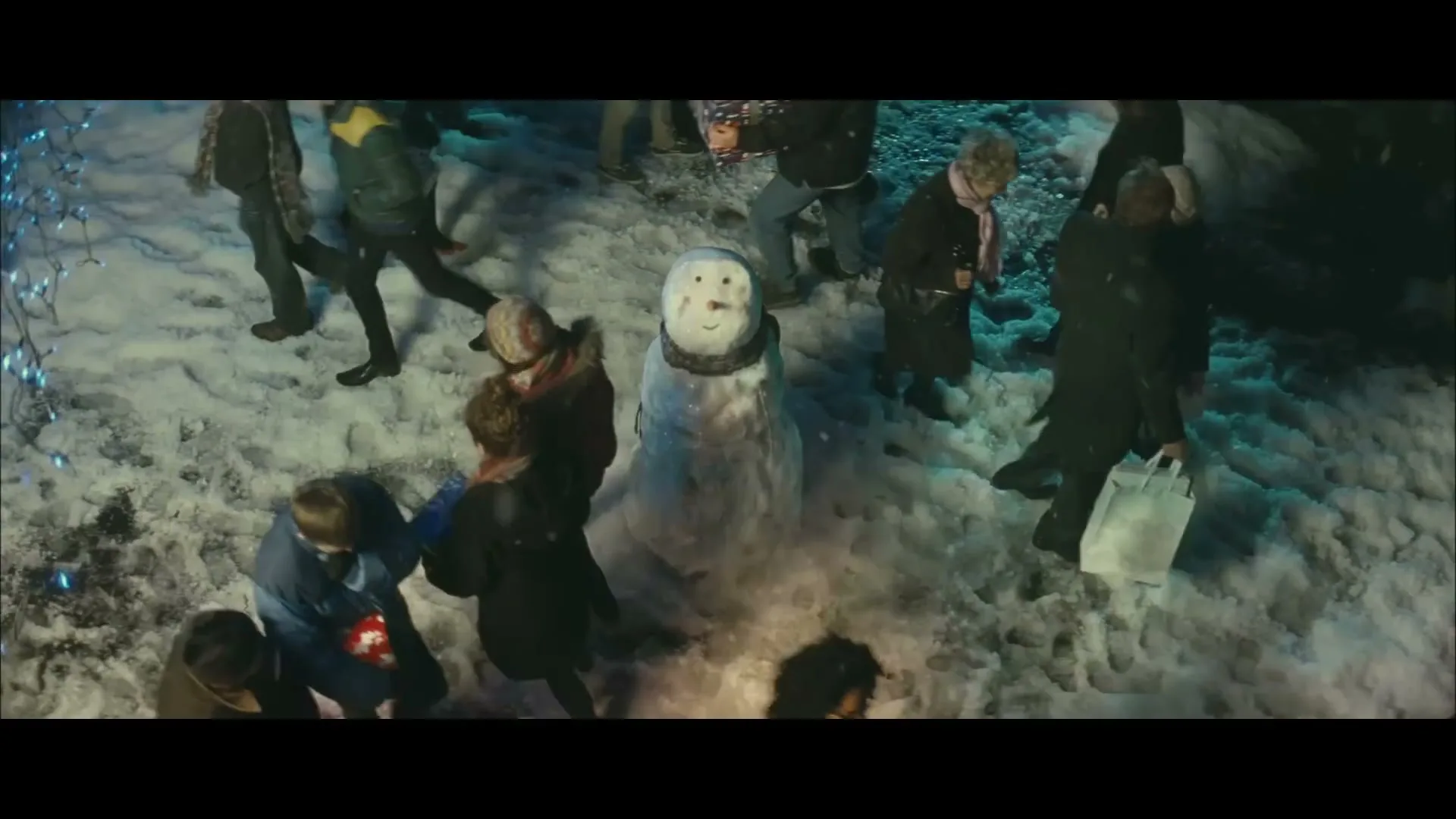 A snowman stands in a snowy, crowded street at night, surrounded by people walking by, some carrying shopping bags, under festive blue lights.