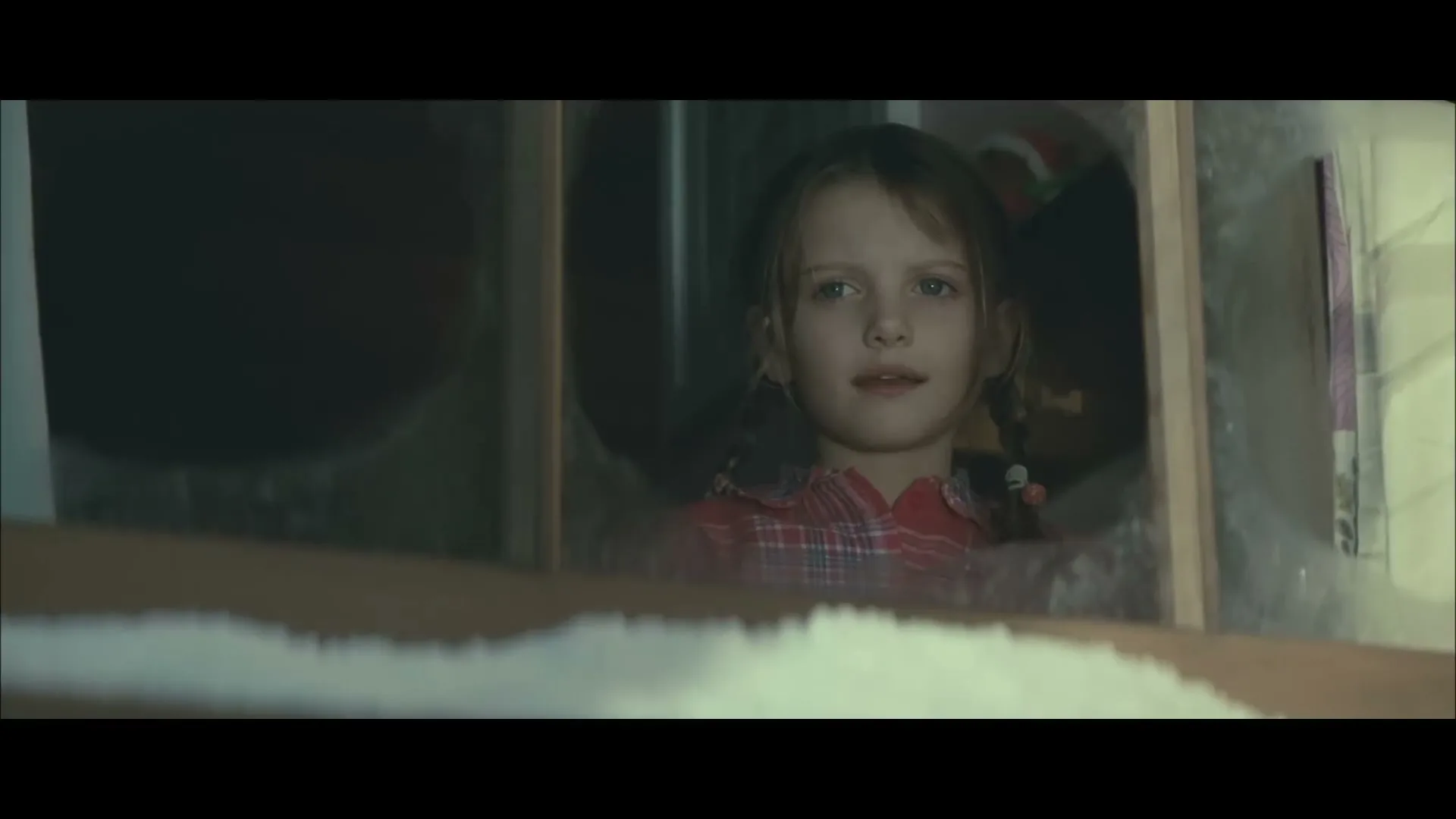 A young girl with pigtails and a red plaid shirt looks out a window, her expression shifting from surprise to a slight smile.