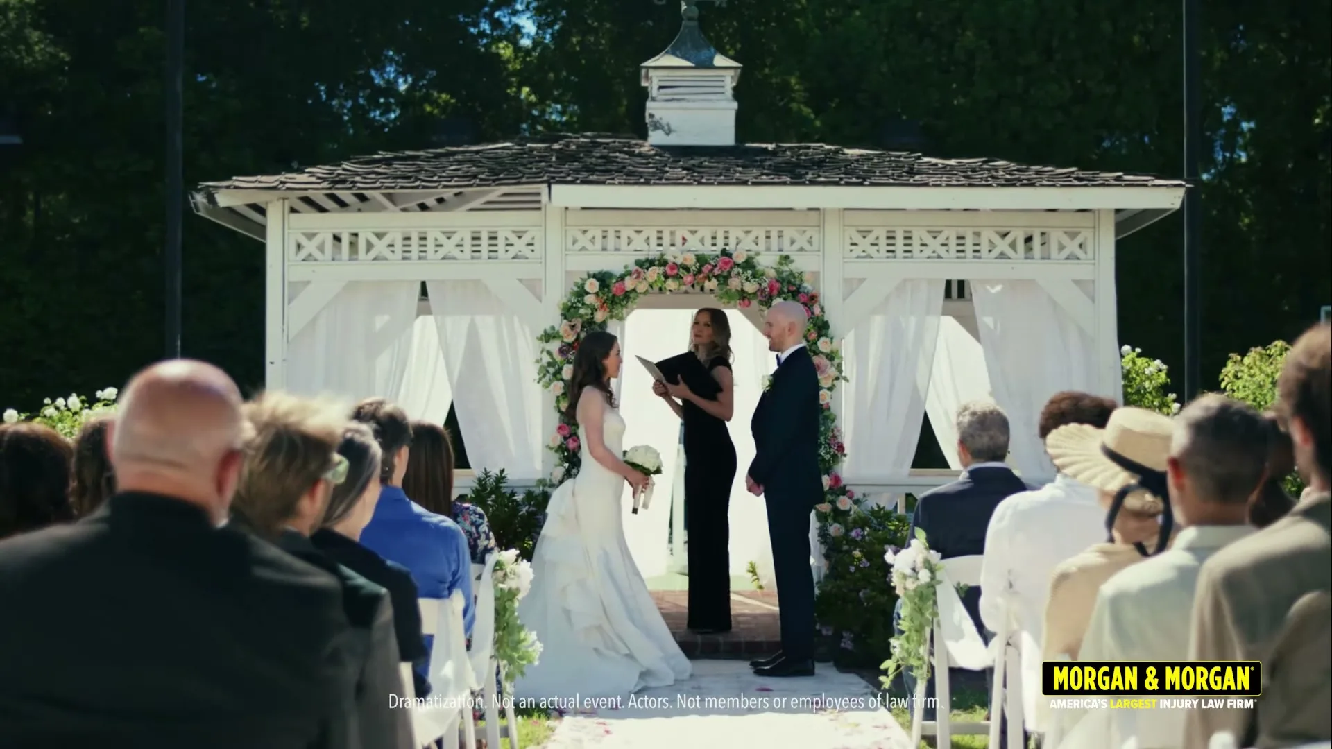 A bride and groom stand at an outdoor wedding ceremony, facing an officiant under a white gazebo adorned with flowers, as guests watch from rows of chairs.