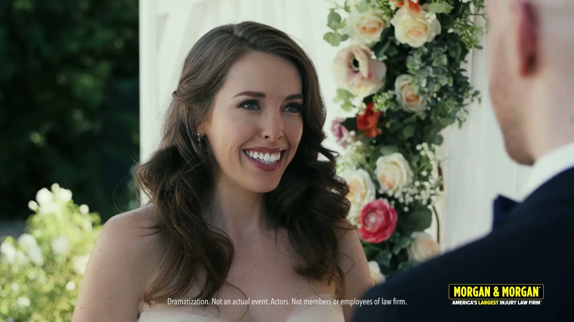A bride in a white dress smiles at the groom during an outdoor wedding ceremony, with floral decorations visible.