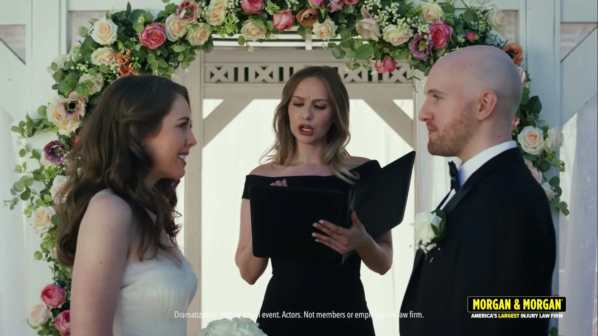 A bride and groom stand at an outdoor wedding altar, while an officiant in a black dress reads from a book, looking concerned.