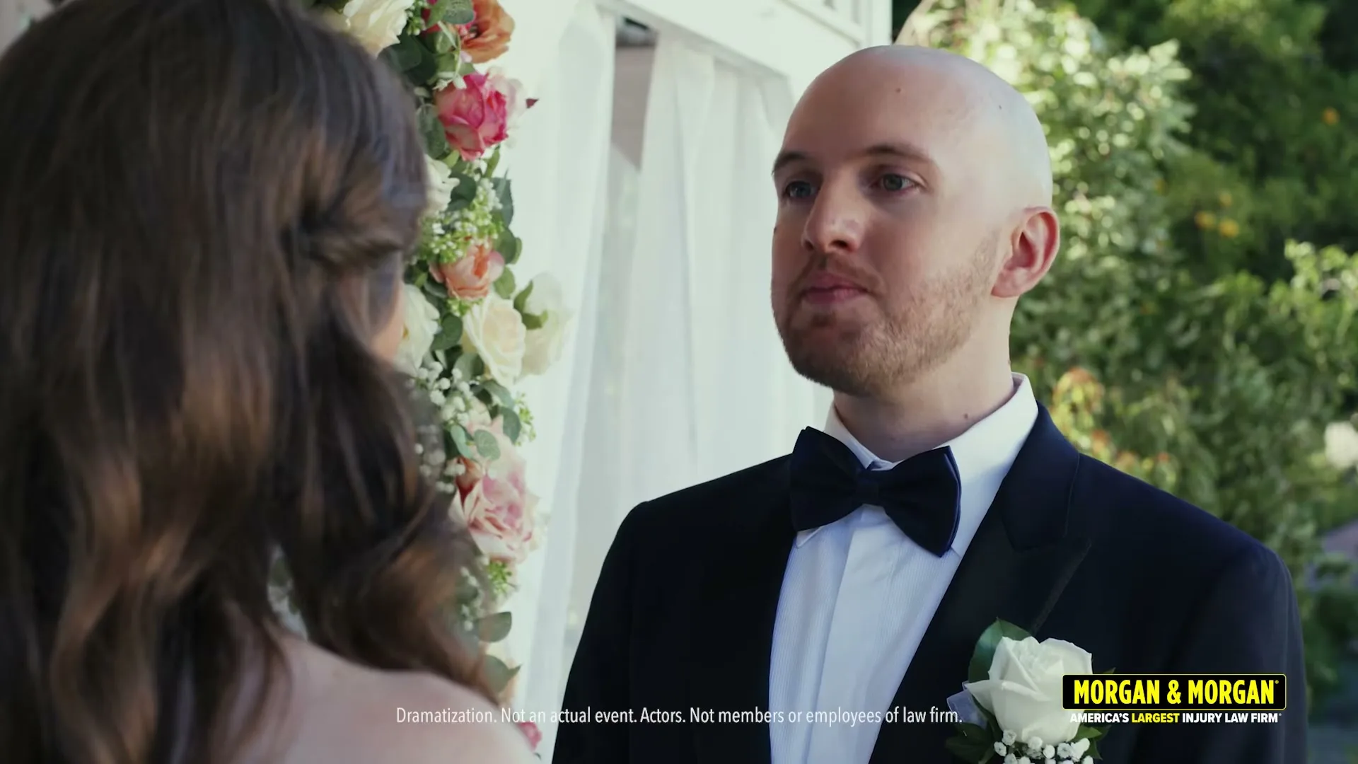 A bald man in a tuxedo with a boutonniere looks at a woman with long brown hair, standing under a floral archway at an outdoor wedding.