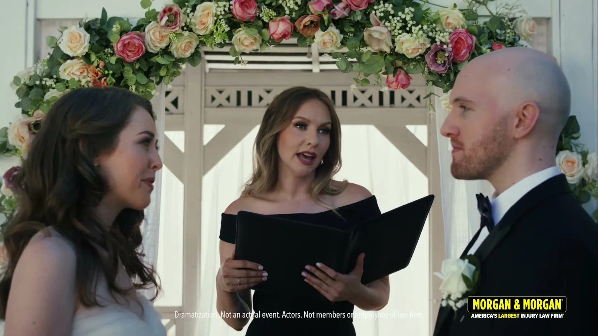 A female officiant in a black dress conducts a wedding ceremony for a bride and groom under a floral archway outdoors.