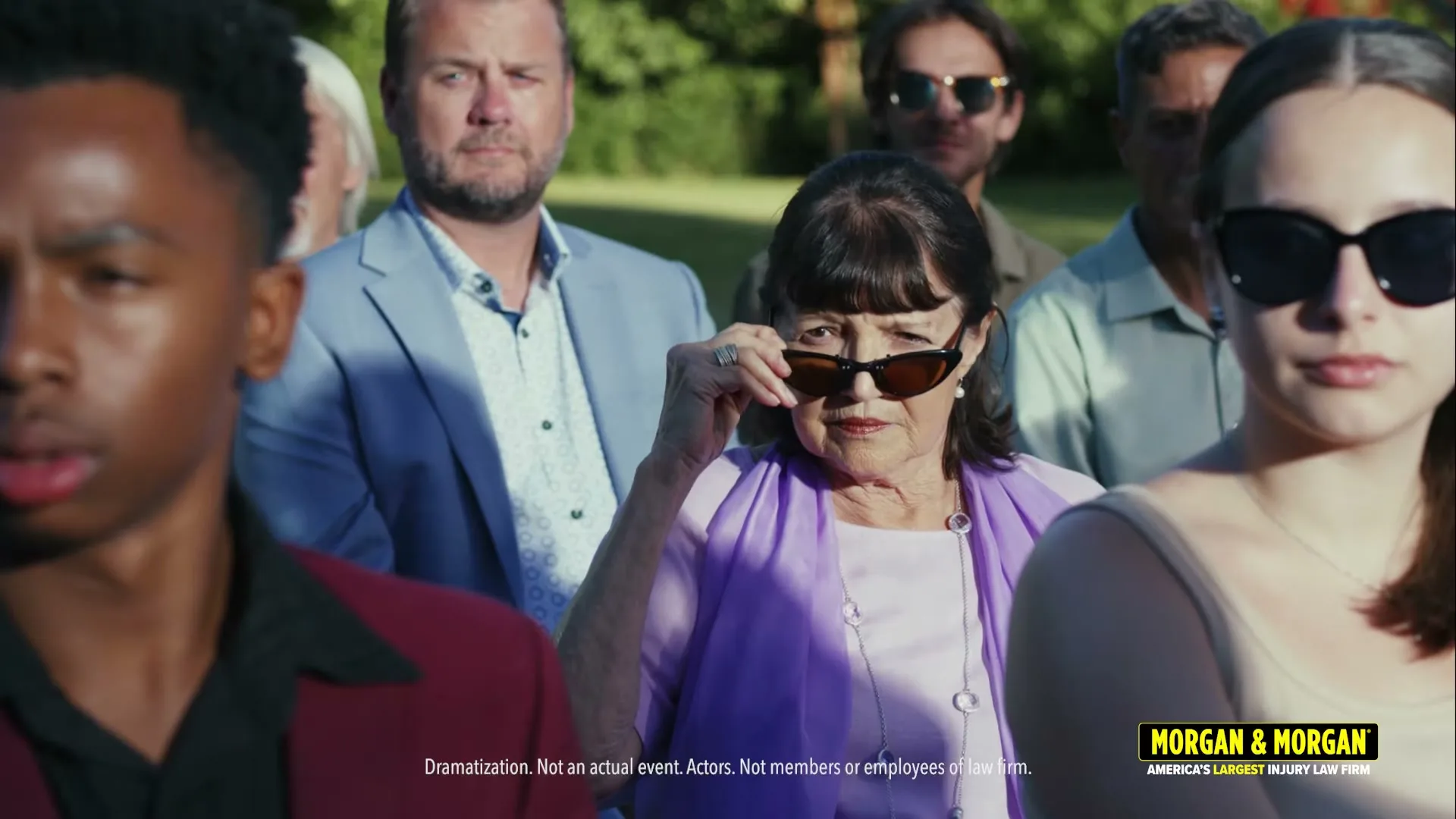 A diverse group of people stand outdoors, looking forward with serious expressions, as an older woman in the center lowers her sunglasses to peer over them.