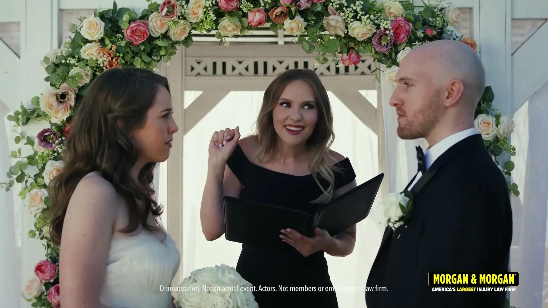 A female officiant in a black dress smiles while holding a book, standing between a bride and groom under a floral archway at an outdoor wedding.