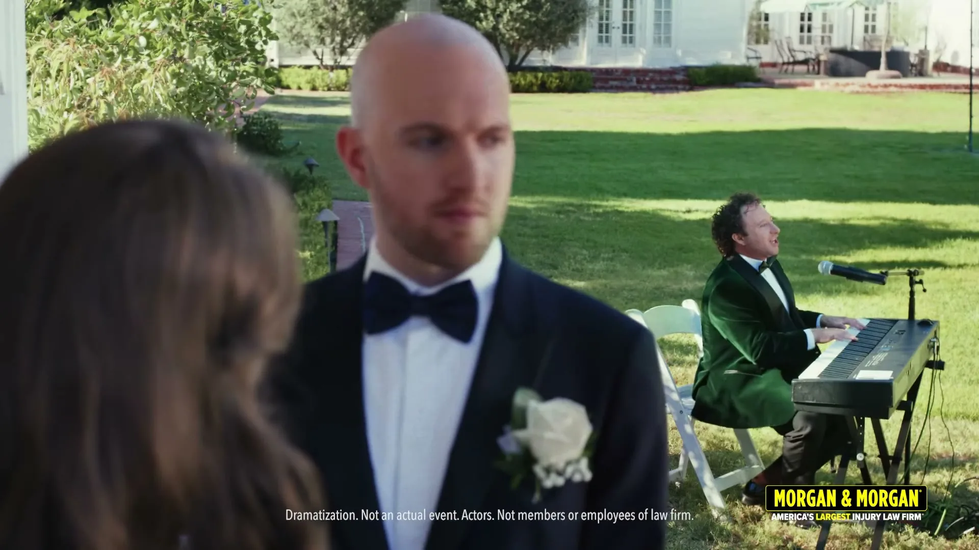 A bald man in a tuxedo with a white rose boutonniere looks to his right, while a man in a green velvet tuxedo plays a keyboard in the background at an outdoor wedding.