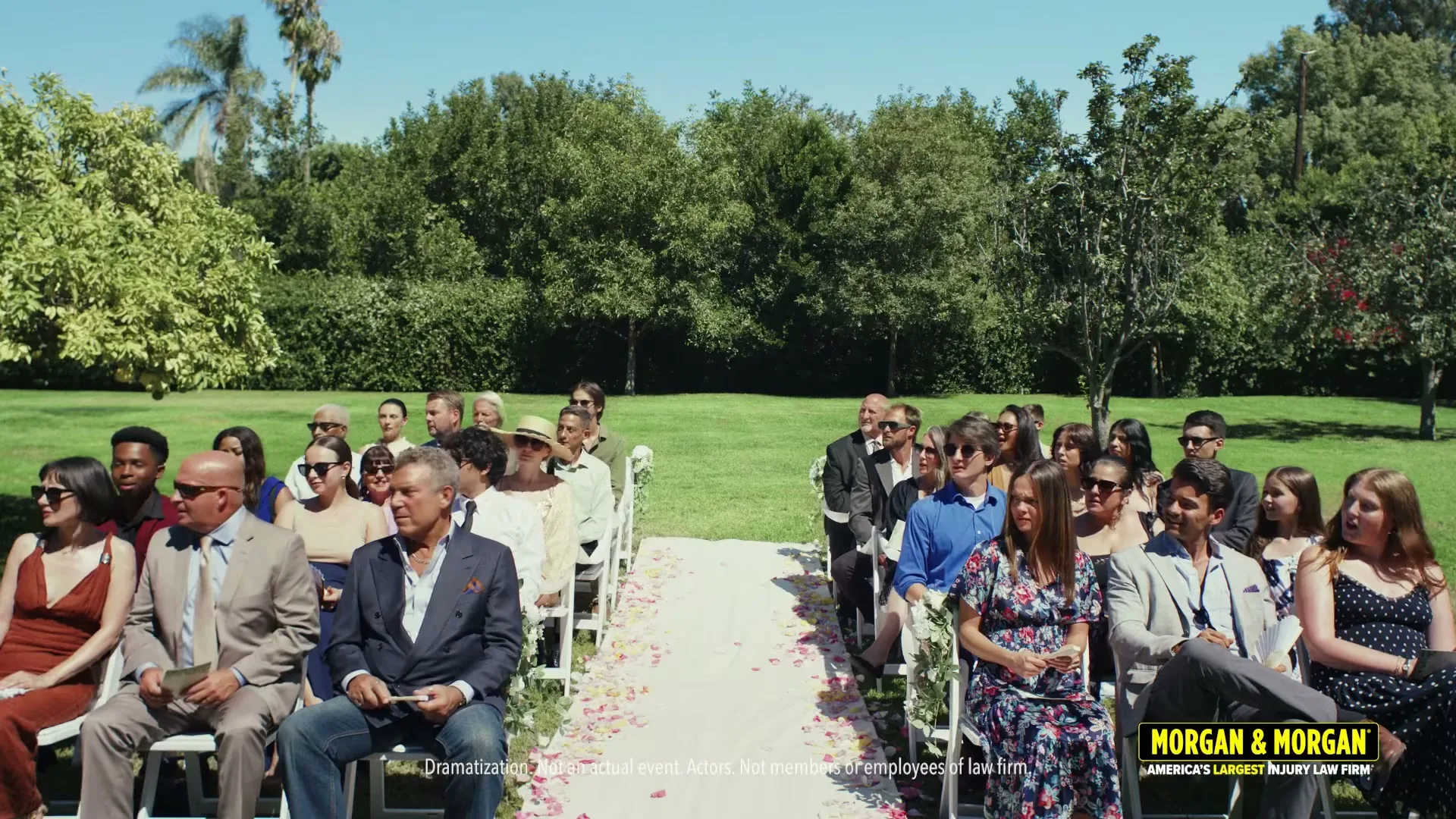 A diverse group of wedding guests, dressed in formal attire, sit in rows of chairs on a sunny green lawn, looking towards the front of the ceremony.