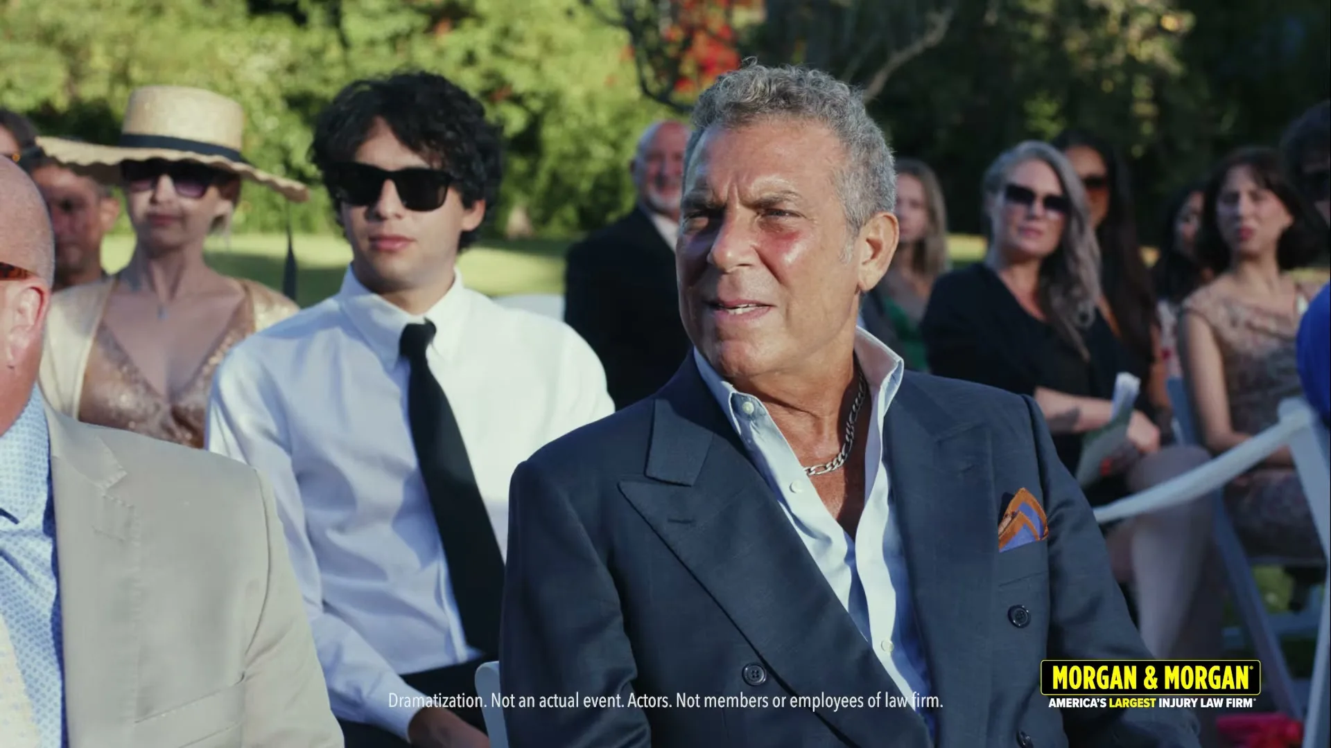 A man in a blue suit with a red mark on his face sits in an outdoor wedding audience, looking to the right.