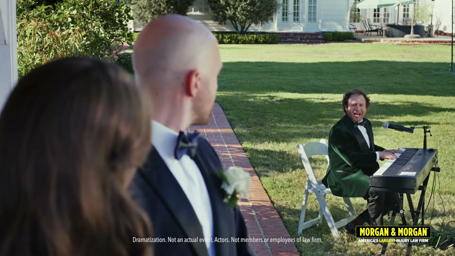 A bald groom in a tuxedo looks over his shoulder at a man in a green velvet tuxedo playing a keyboard and singing at a wedding.