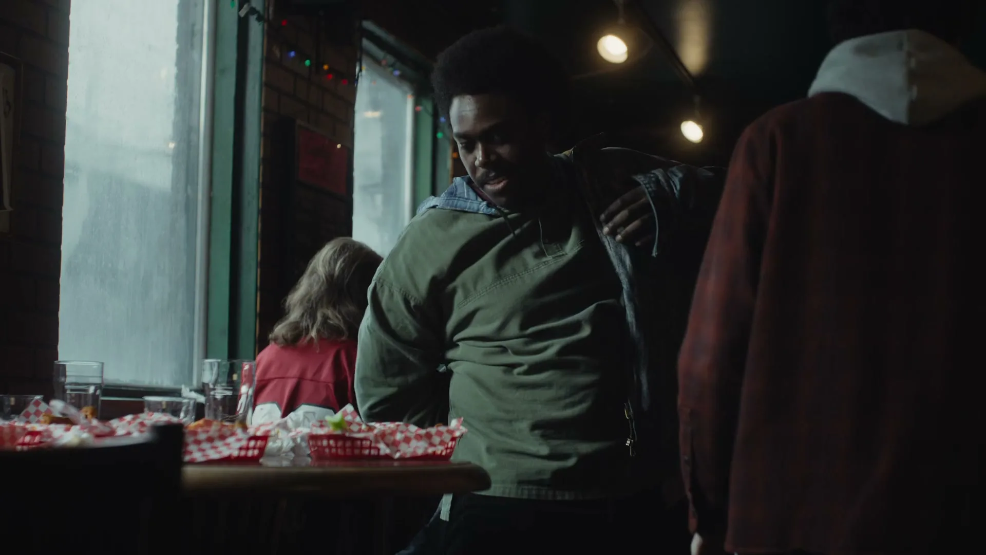 A young Black man in a green jacket walks past a table with food baskets in a dimly lit restaurant, while other patrons are visible in the background.