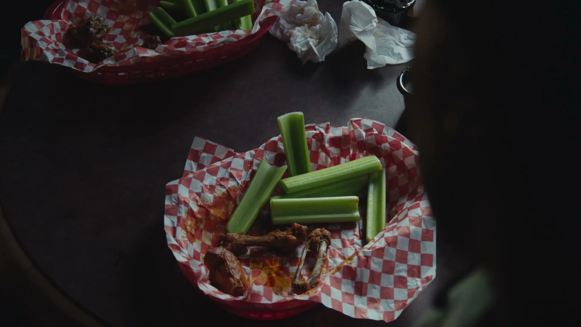 A high-angle shot shows two red checkered baskets of celery sticks and chicken wing bones on a dark wooden table in a dimly lit restaurant.