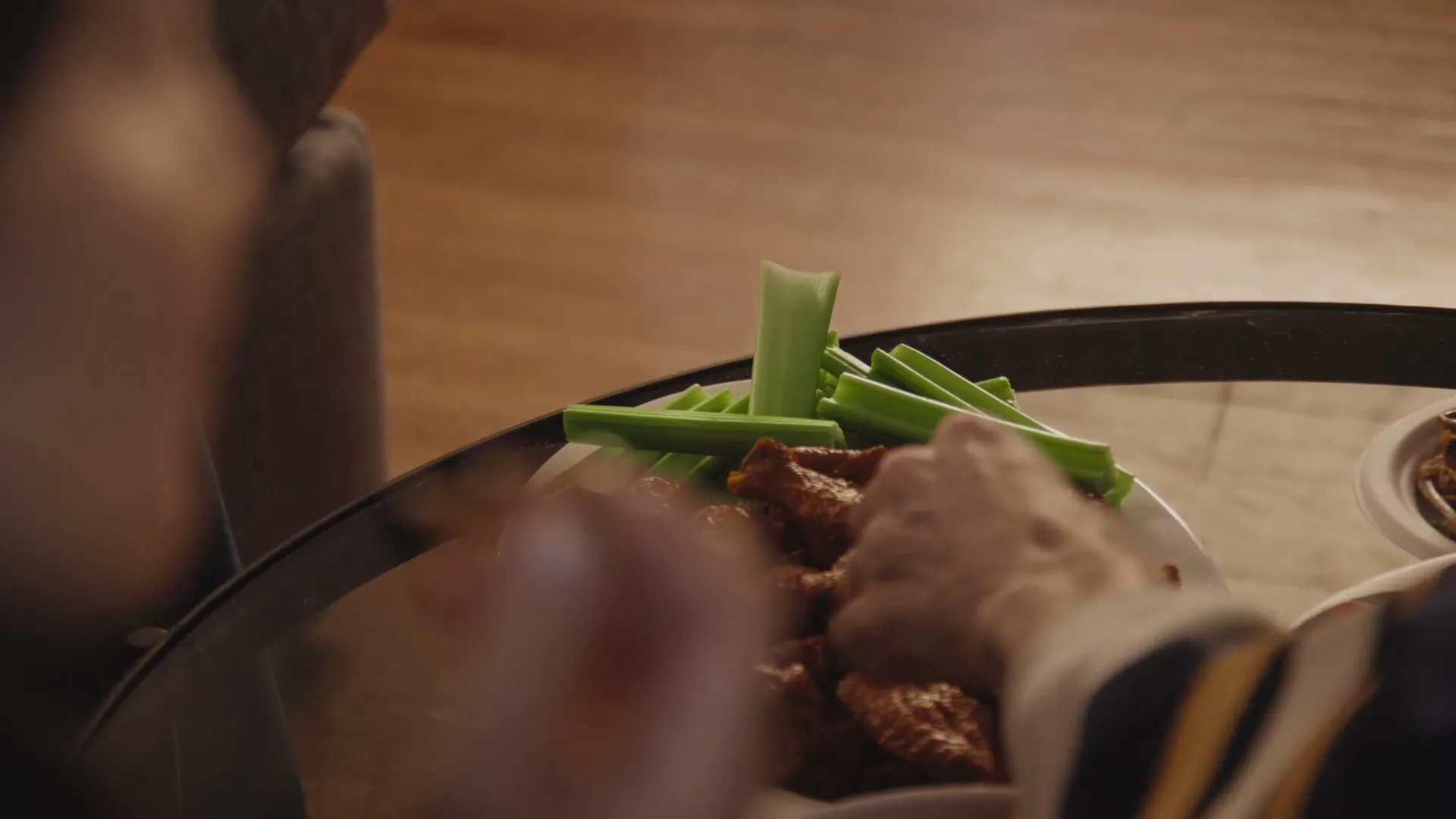 A person's hand reaches for a chicken wing from a plate of wings and celery sticks on a glass coffee table in a living room.