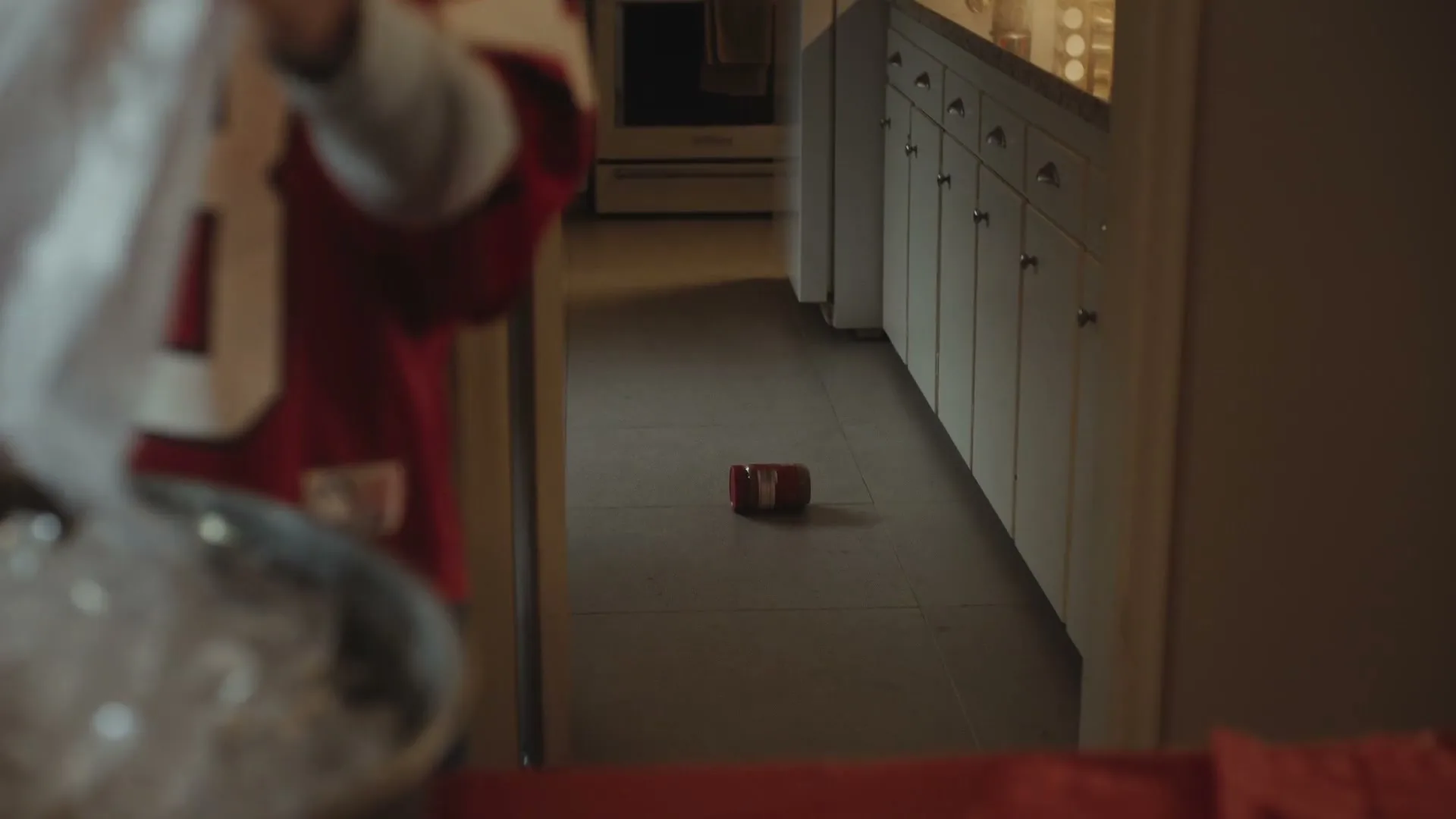 A child in a red jersey is out of focus in the foreground, while a small red Jif peanut butter container lies on the floor in a kitchen hallway.