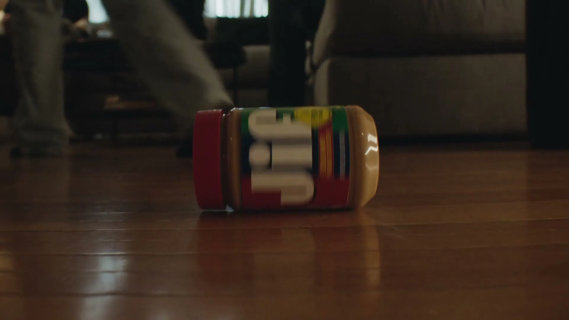A jar of Jif peanut butter rolls across a polished wooden floor in a dimly lit living room.